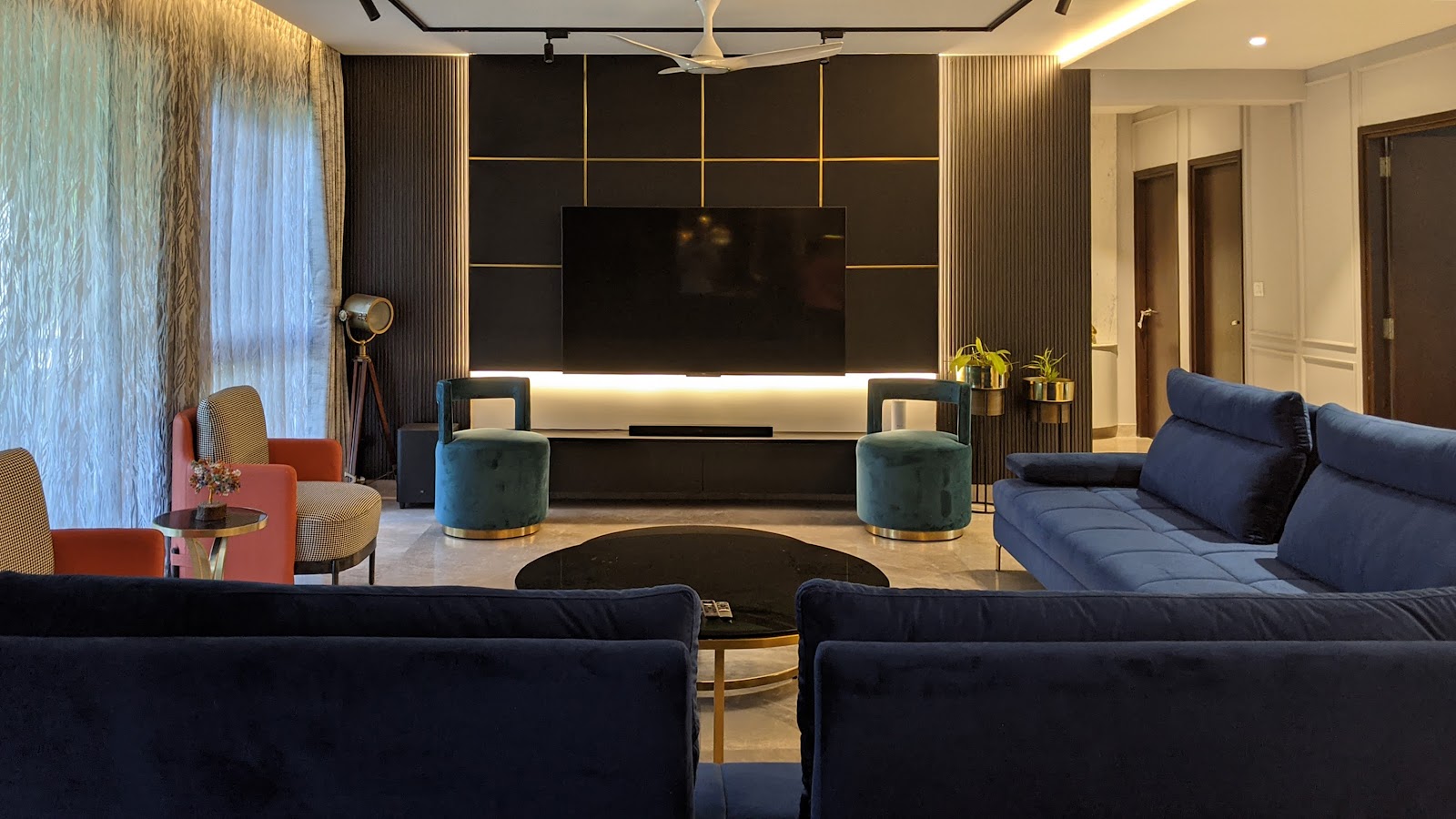 Living room with a black and brass grid-panel TV wall, a navy velvet sectional sofa, emerald velvet stools, a rust-orange armchair