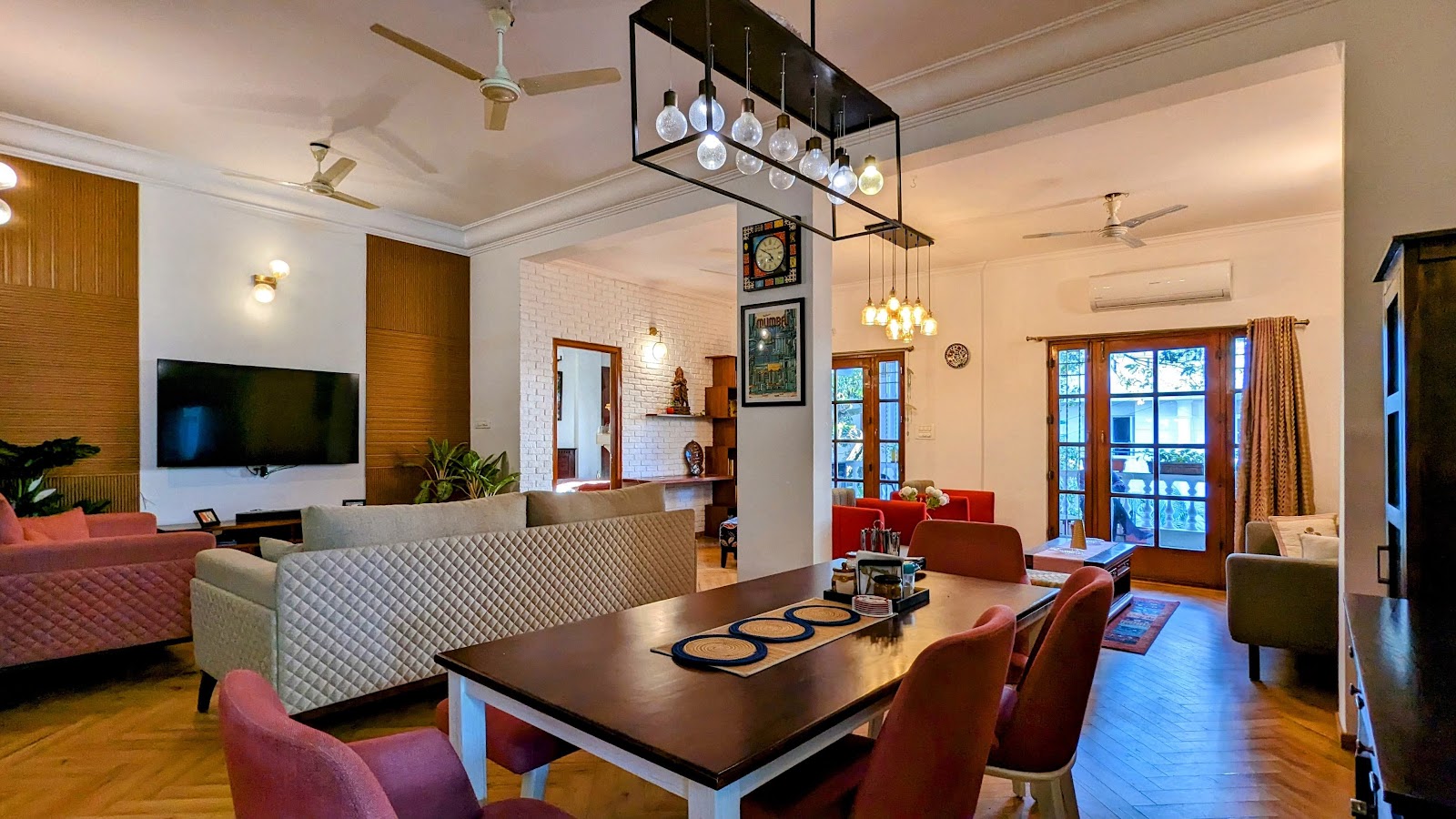 Open-plan living-dining space with a quilted grey sofa, mismatched pink and red dining chairs, a cage-style multi-bulb pendant