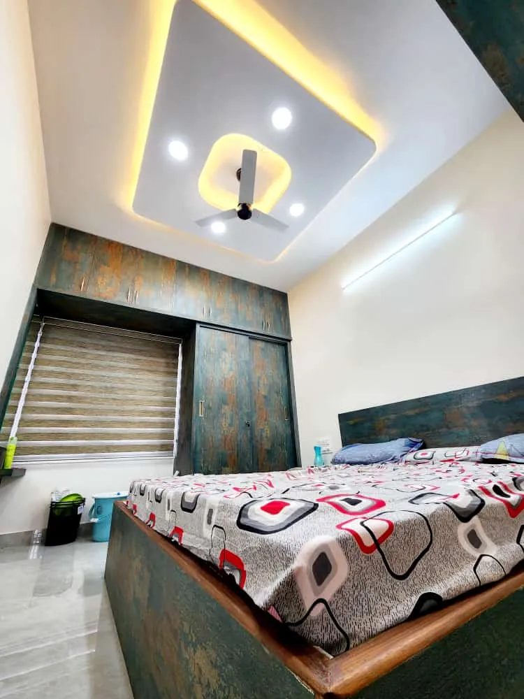 Photograph taken from the bed corner of a bedroom whose teal-and-rust distressed laminate wardrobe with sliding shutters and loft flanks a zebra-blinded