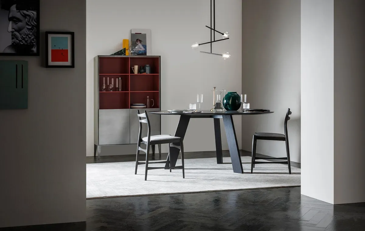 Round ebonised dining table on a faceted trestle base with two ladder-back chairs, set on a pale rug over dark herringbone parquet