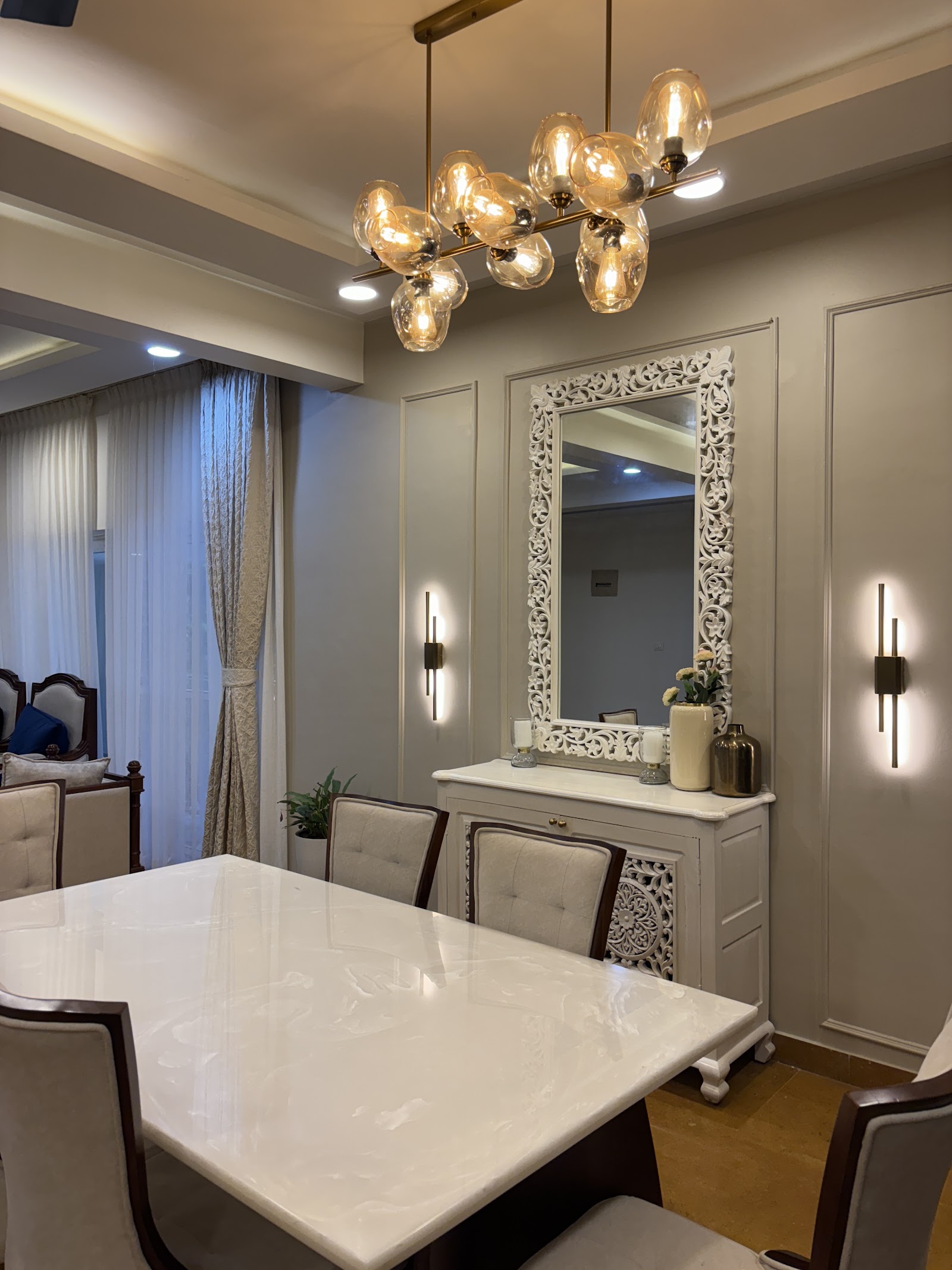 Dining area with a white marble-top table, upholstered dining chairs, an ornate carved mirror over a console, and a cluster glass-globe chandelier.