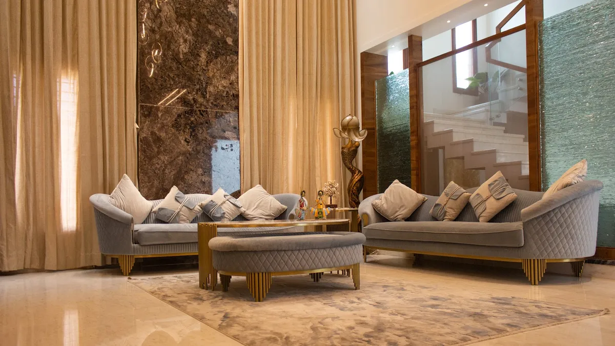 Double-height living room where two grey quilted-velvet sofas on fluted gold plinths face a book-matched brown marble slab panel between full-height beige