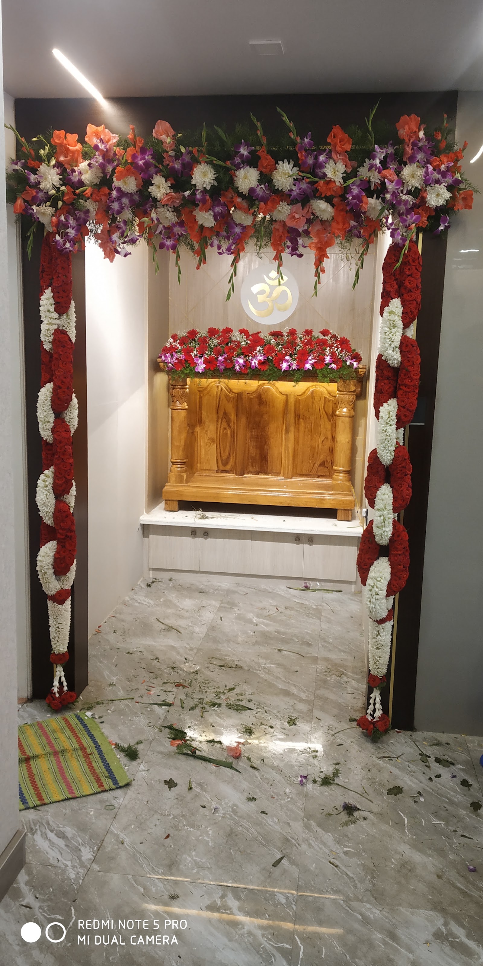 Pooja room with a carved wooden mandir unit and a backlit Om symbol, framed by fresh floral garlands and petals from a housewarming ceremony.