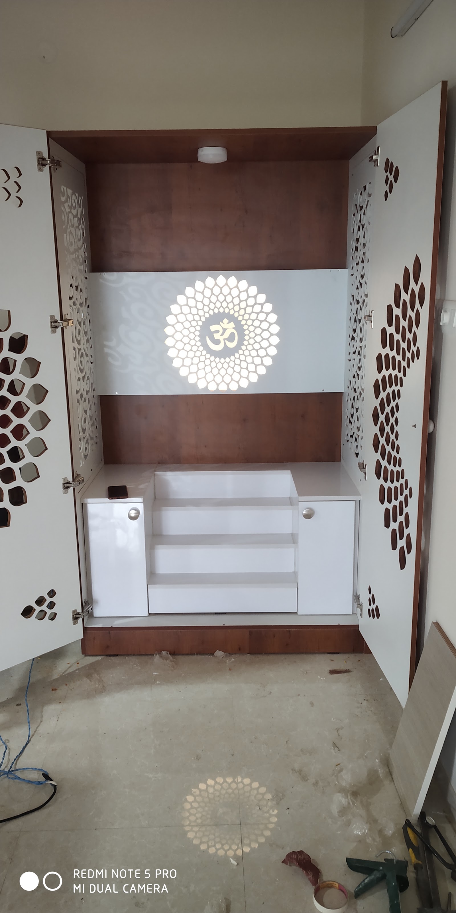 Newly installed white pooja cabinet with jaali-cut cutwork doors and a backlit Om lotus panel, floor still dusty with tools from installation.