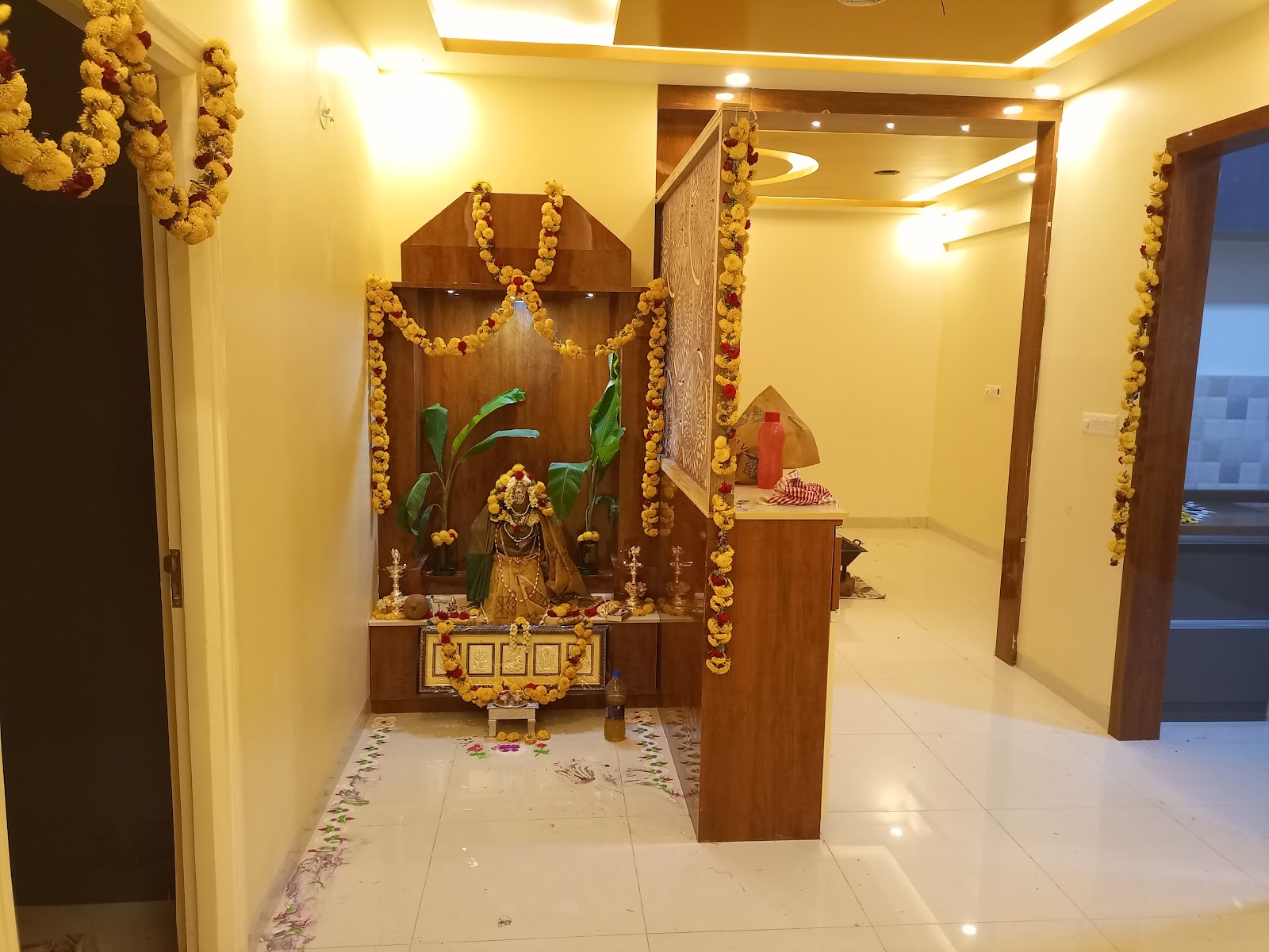 Home pooja unit with a carved wood mandir housing a garlanded deity idol, flanked by a jaali-carved wood partition, set in a foyer with cove-lit ceiling.