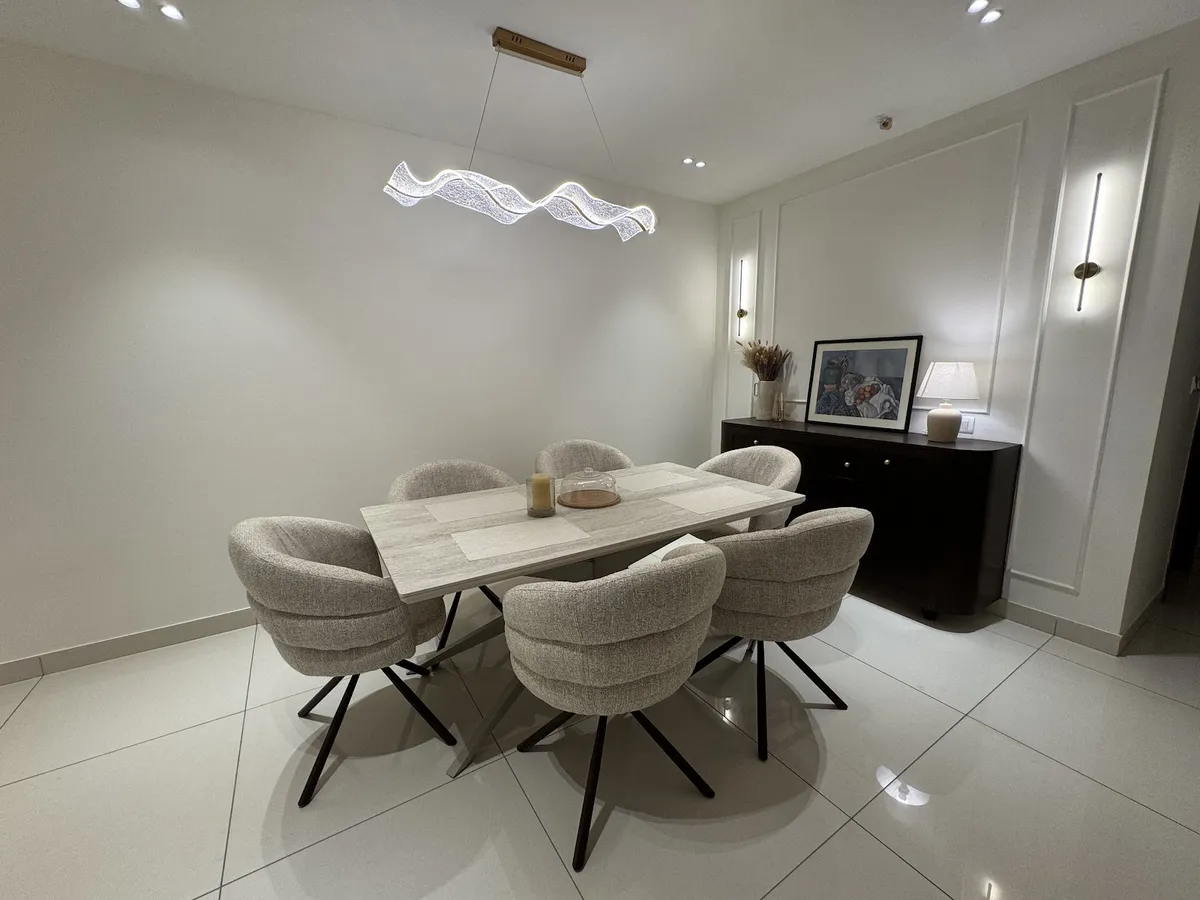 A dining room with a travertine-top table, six boucle swivel chairs, a sculptural wavy LED pendant light, and a dark wood console styled with framed art.