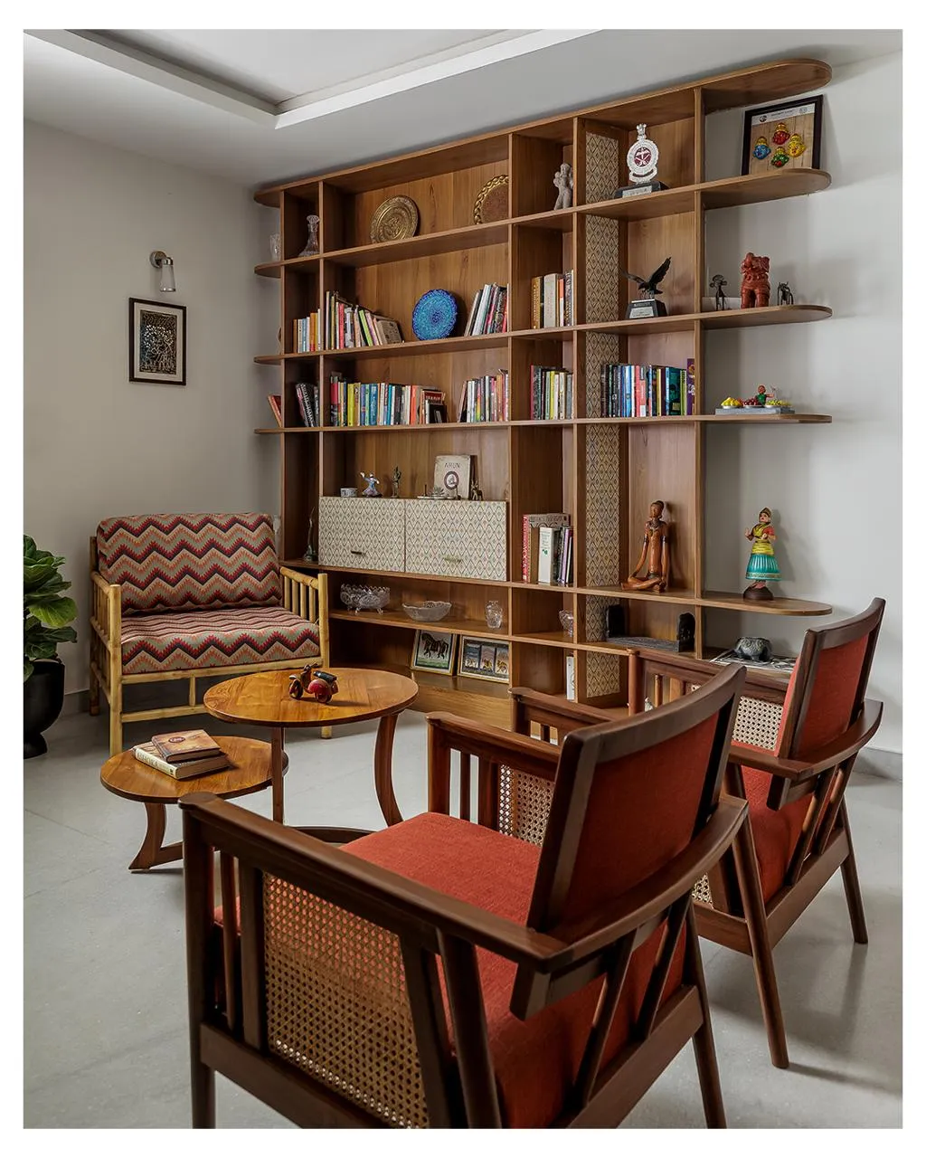 Reading corner where a floor-to-ceiling teak shelf of books, brass plates and terracotta figures faces two cane-backed teak armchairs in rust wool and an