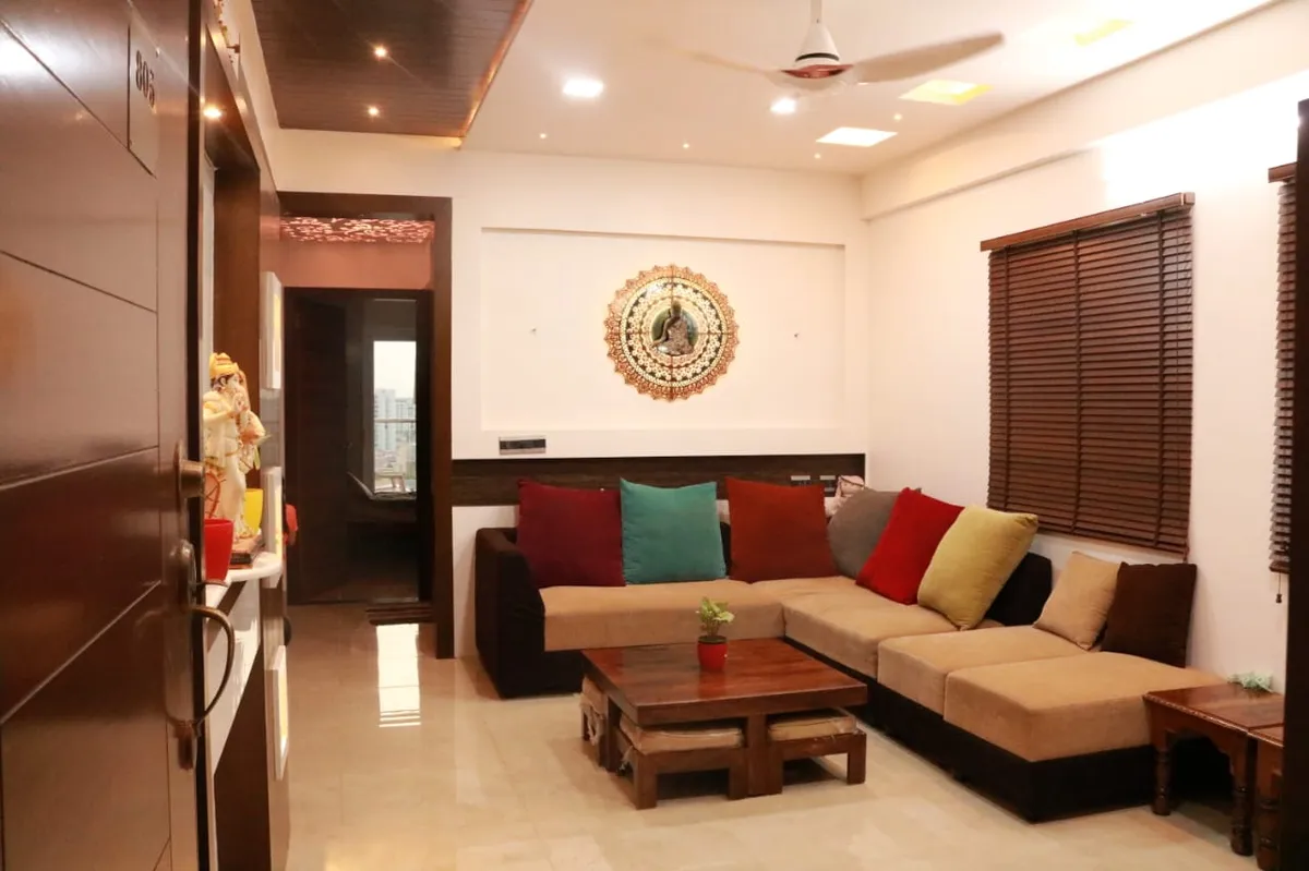 Apartment living room seen from the open entrance door numbered 805: beige-and-brown L-sofa with red, teal and mustard cushions