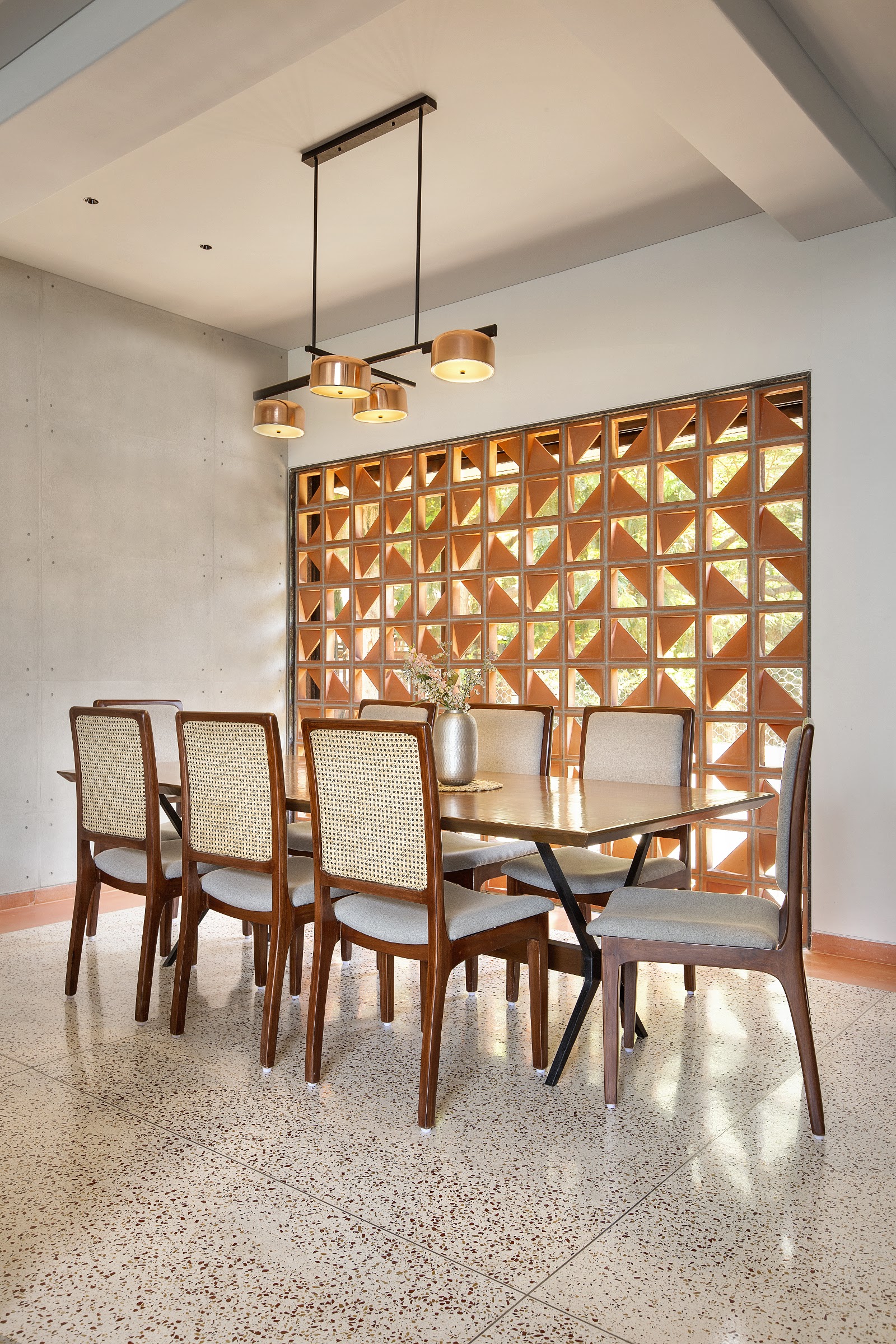 Dining room with a terracotta jaali screen wall and cane-back wood dining chairs around a table, lit by a cluster copper pendant light.