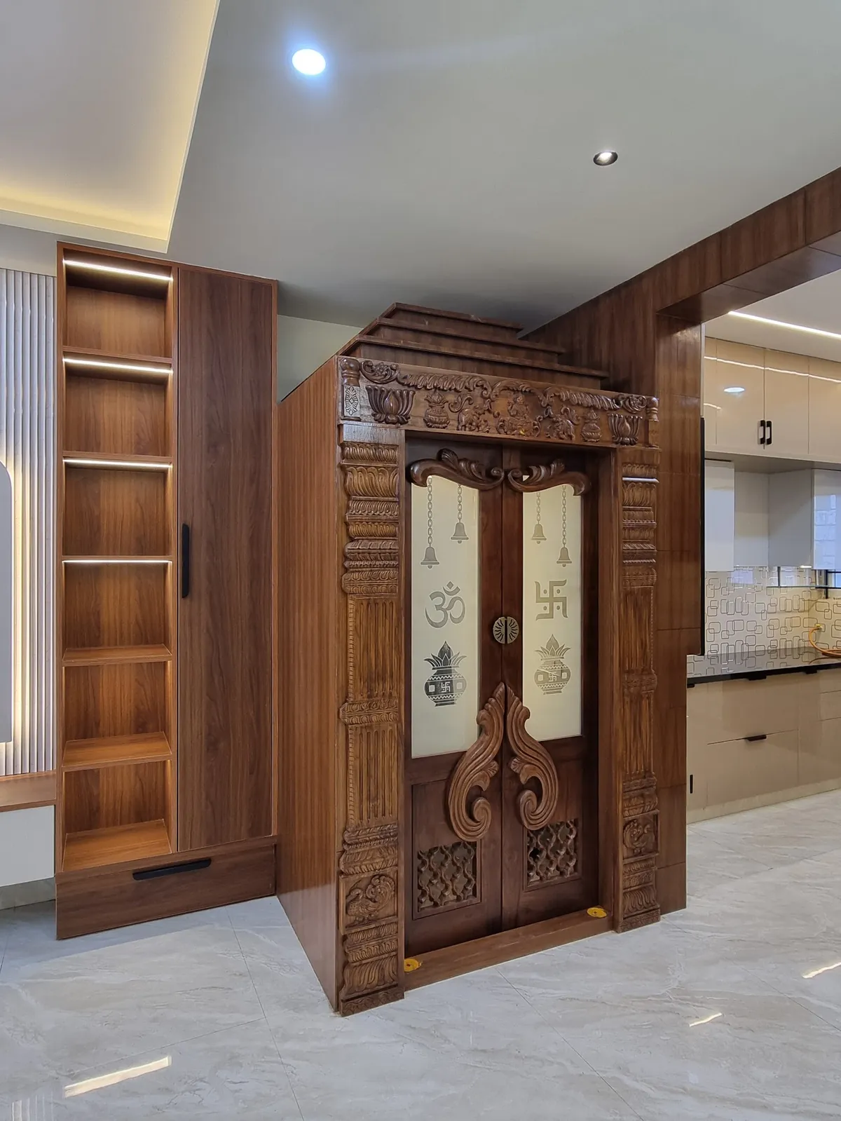 Carved wooden pooja unit with ornately detailed double doors featuring Om and Swastik motifs and hanging bells, flanked by an open wood shelving column