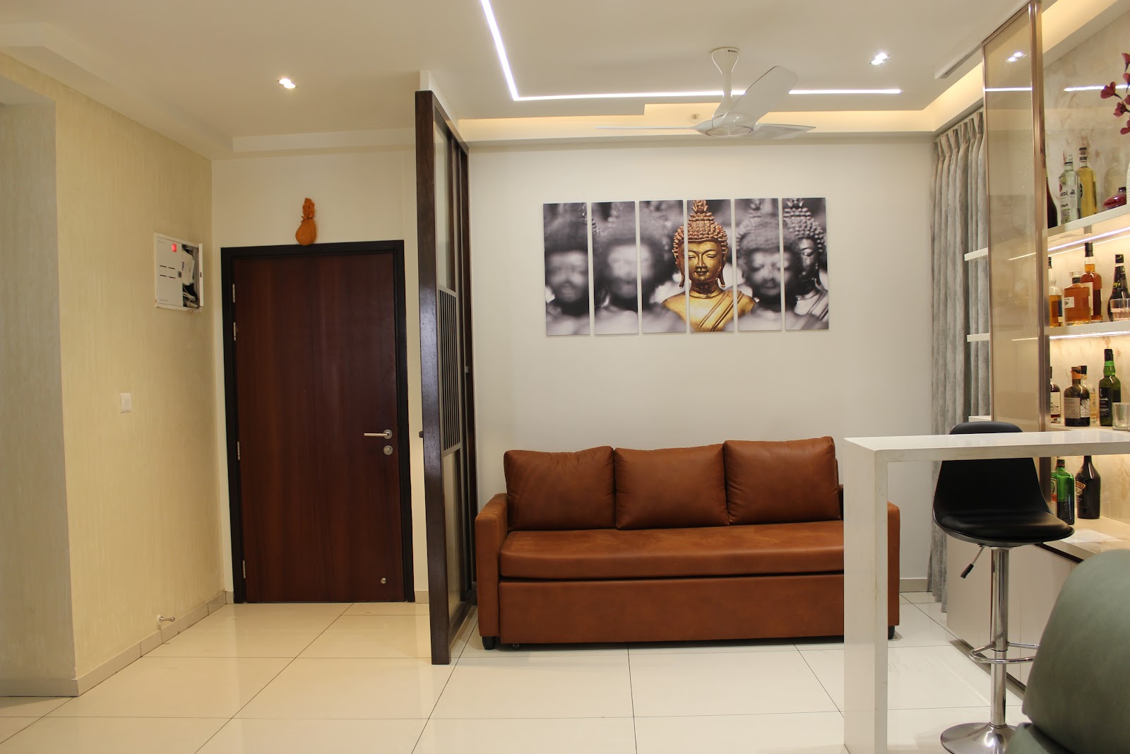 Entrance hallway opening into a living area with a tan leather sofa beneath a six-panel Buddha photographic wall art, cove-lit ceiling