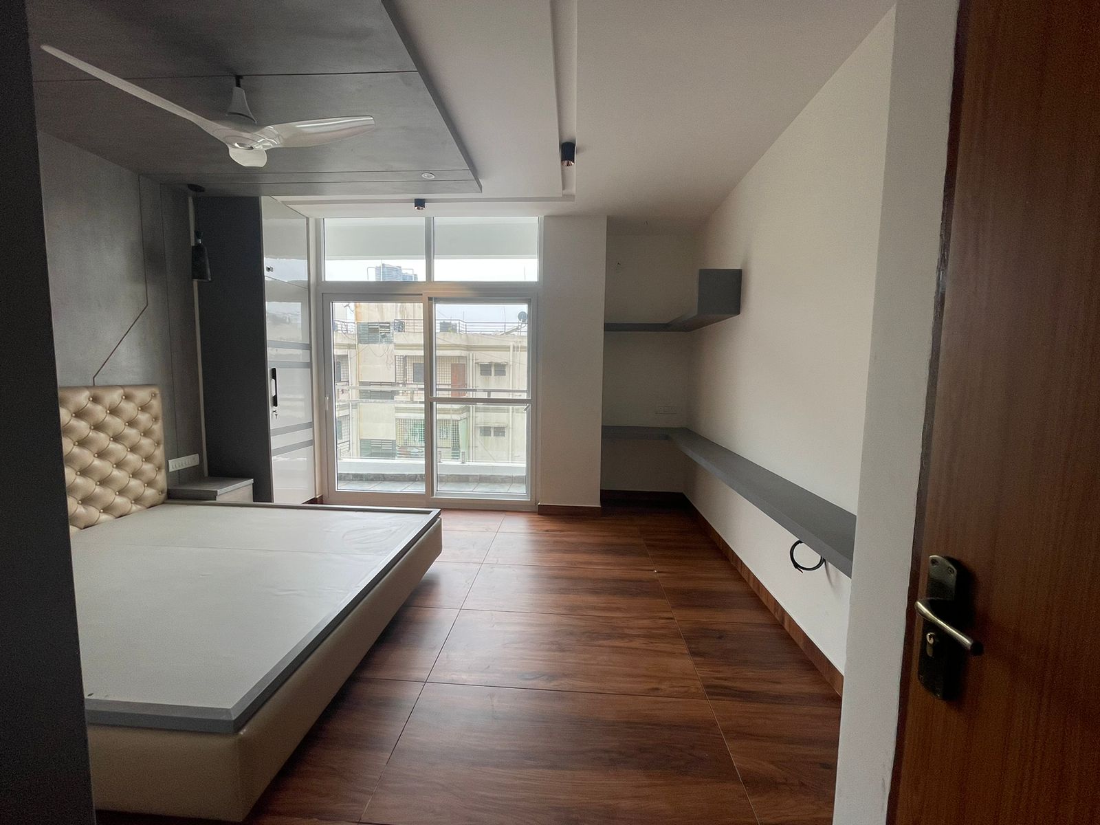 Bedroom with a champagne tufted platform bed, wood-look tile flooring and a plain grey ledge shelf leading to a balcony.