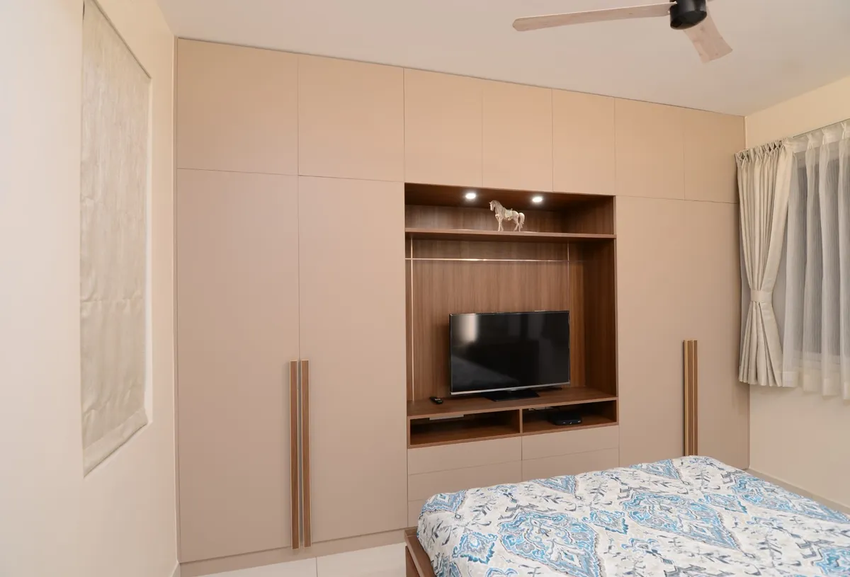 Bedroom wall built out as a floor-to-ceiling beige wardrobe run with lofts, wrapping a walnut-veneer TV niche lit by two small spots with an open drawer