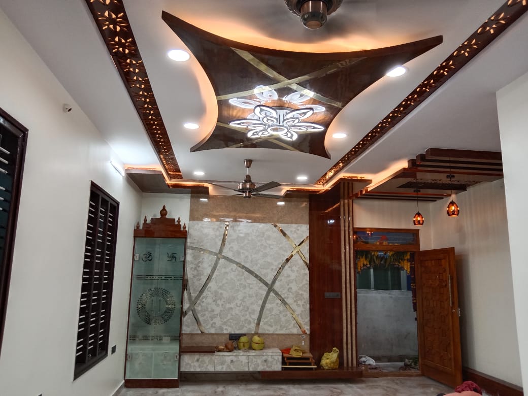 Entrance foyer with a sculpted wing-shaped POP false ceiling with jaali trim, a carved wood main door, and a glass-door pooja cabinet in the corner.