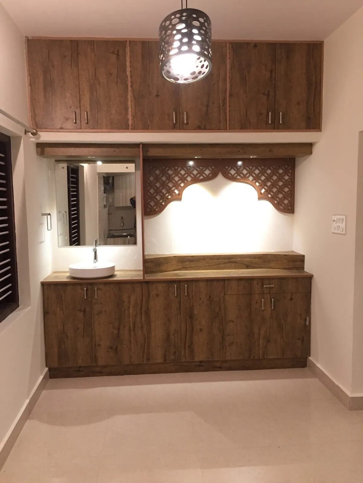 Rustic-oak laminate pooja unit with a CNC-carved arched jaali pelmet over a backlit white niche