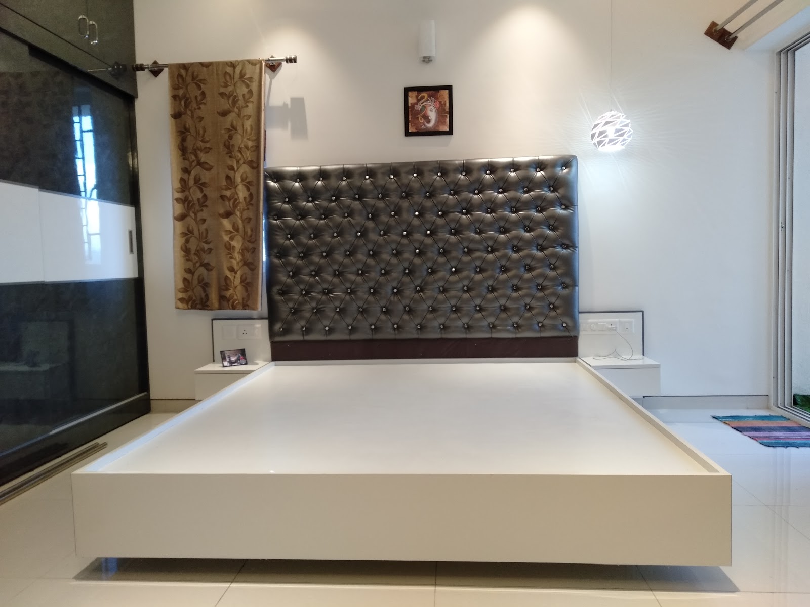 Bedroom with a crystal-studded silver tufted headboard, a floating white platform bed base, and a dark glass-shutter wardrobe beside a geometric pendant light.