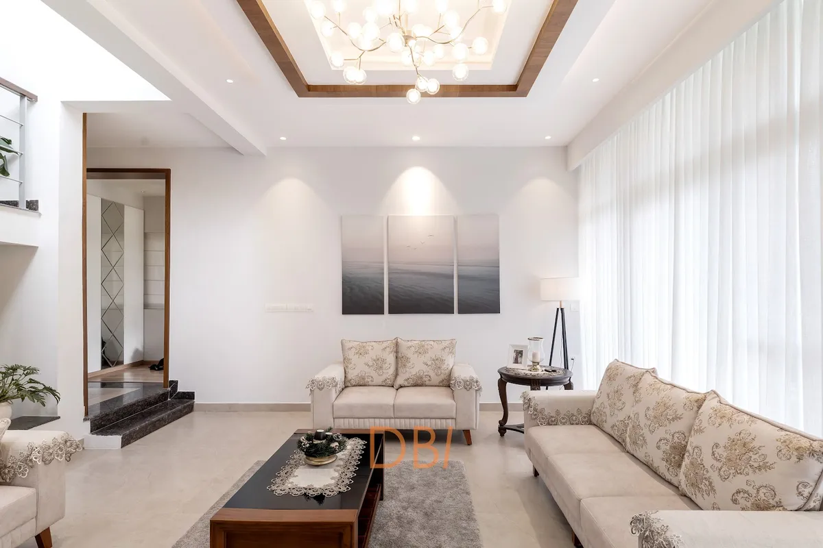 Double-height living room with a sculptural branch chandelier set into a wood-trimmed cove ceiling, floral lace-trimmed cream sofas, a black wood coffee table