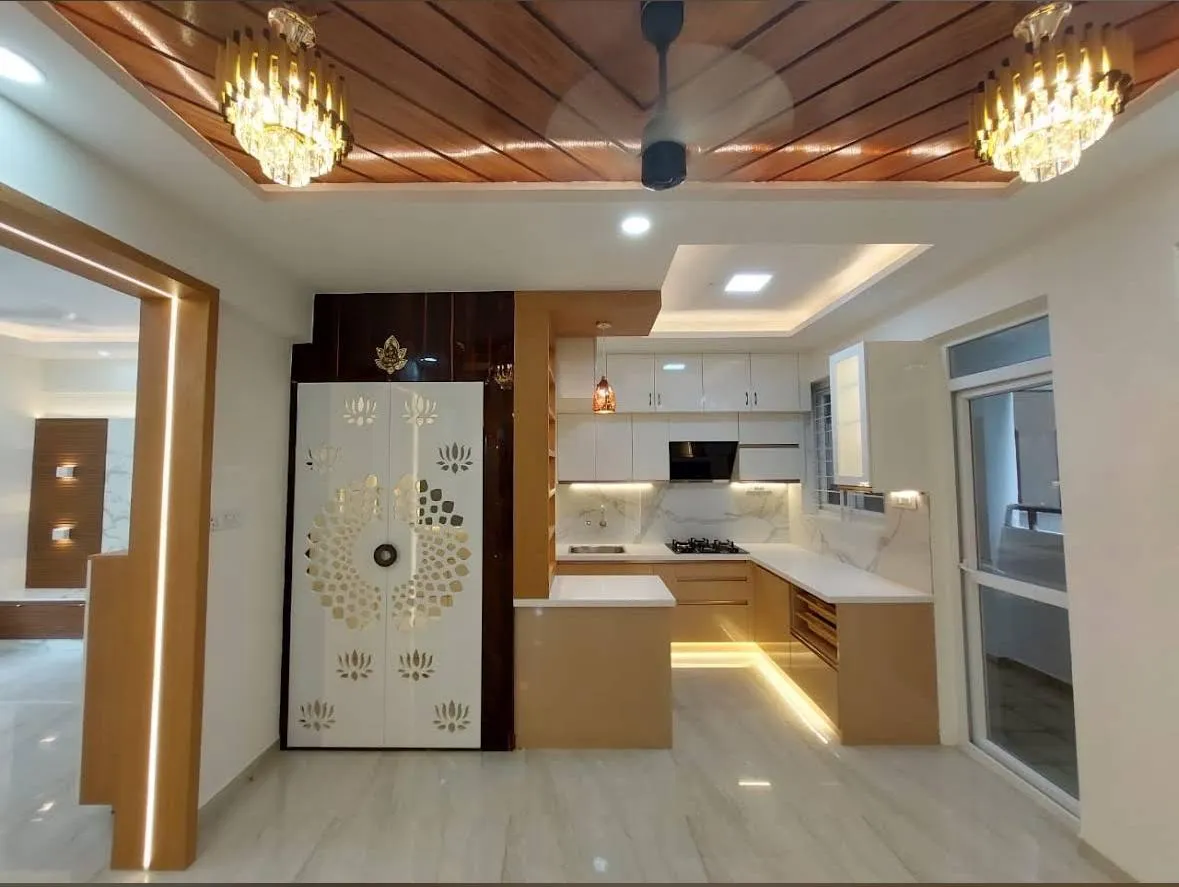 Open-plan frame with a gold lotus-jaali pooja unit standing between a beige-and-white U-shaped kitchen and the living area