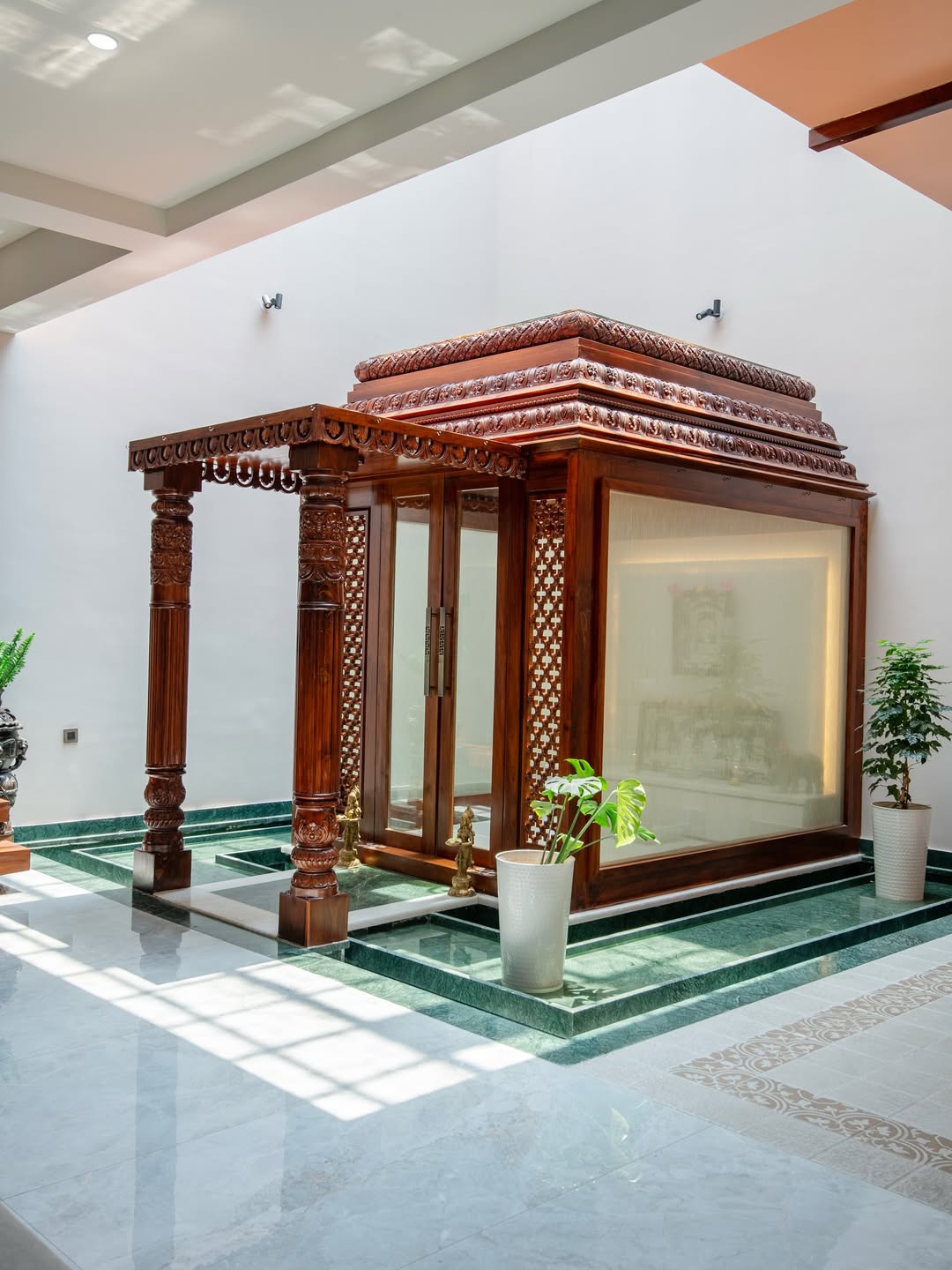 Freestanding carved teak pooja mandir with jaali lattice side panels, glass doors, and a raised green marble platform in a double-height foyer.