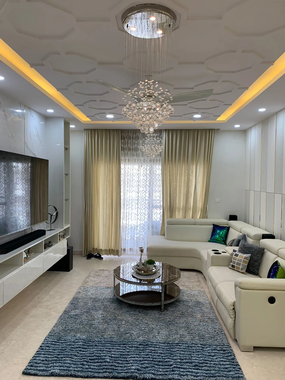 Cream leather recliner sofa and a round smoked-glass table on a blue shag rug, under a honeycomb-relief POP ceiling where a crystal chandelier hangs inside the
