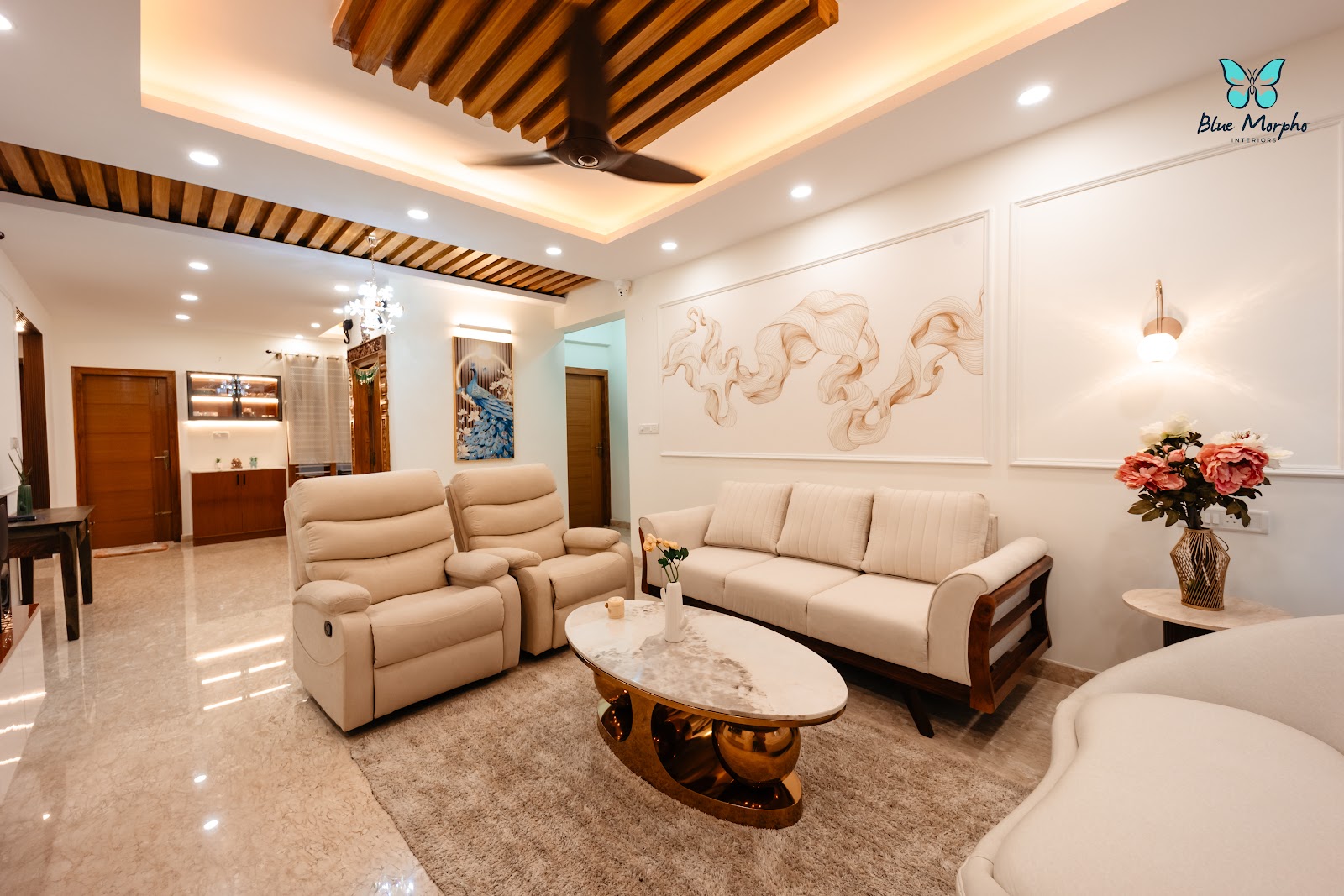 Living room with a wood-slat coved false ceiling, cream reclining sofa set, and an oval marble-and-gold coffee table on a beige shag rug.