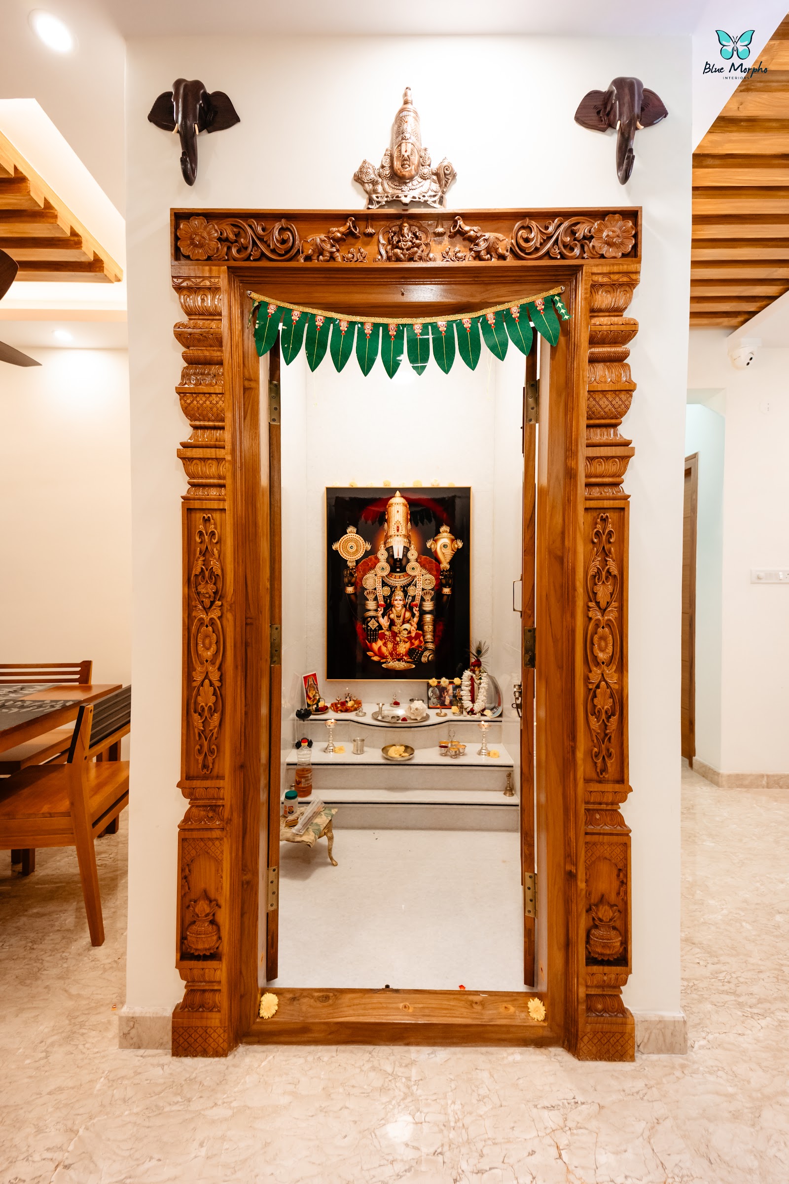 Dedicated pooja room with an elaborately hand-carved teak door frame with elephant and floral motifs, a mango-leaf toran