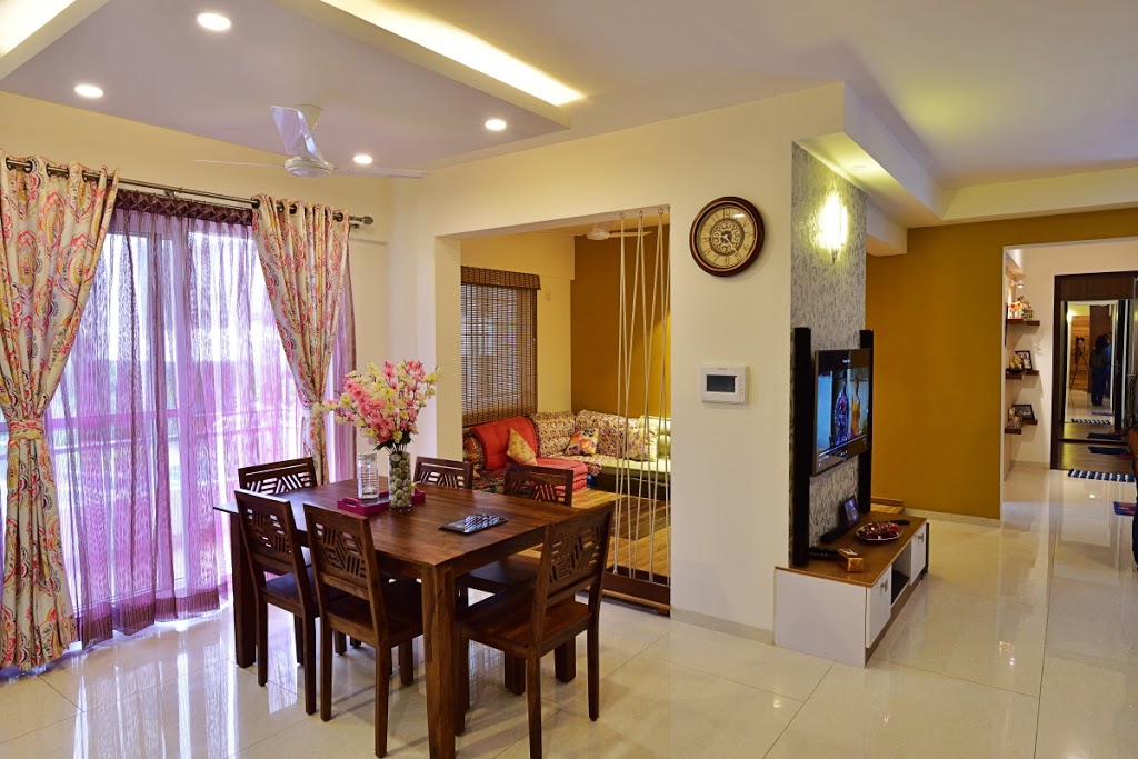 Open dining-and-living space with a wood dining table under pendant lighting, a TV wall in mustard paint, and a floor-seating swing nook beyond.