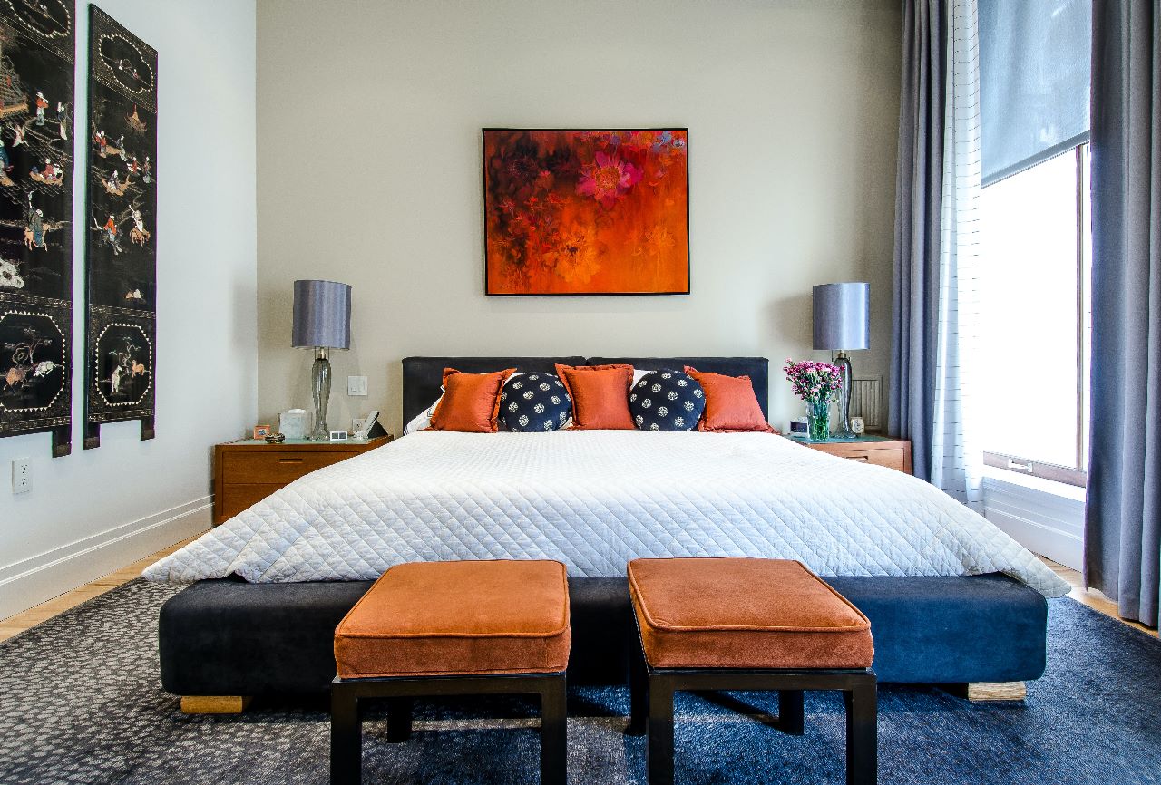 Bedroom with a navy upholstered bed dressed in orange and navy accent pillows, a fiery abstract floral painting above the headboard