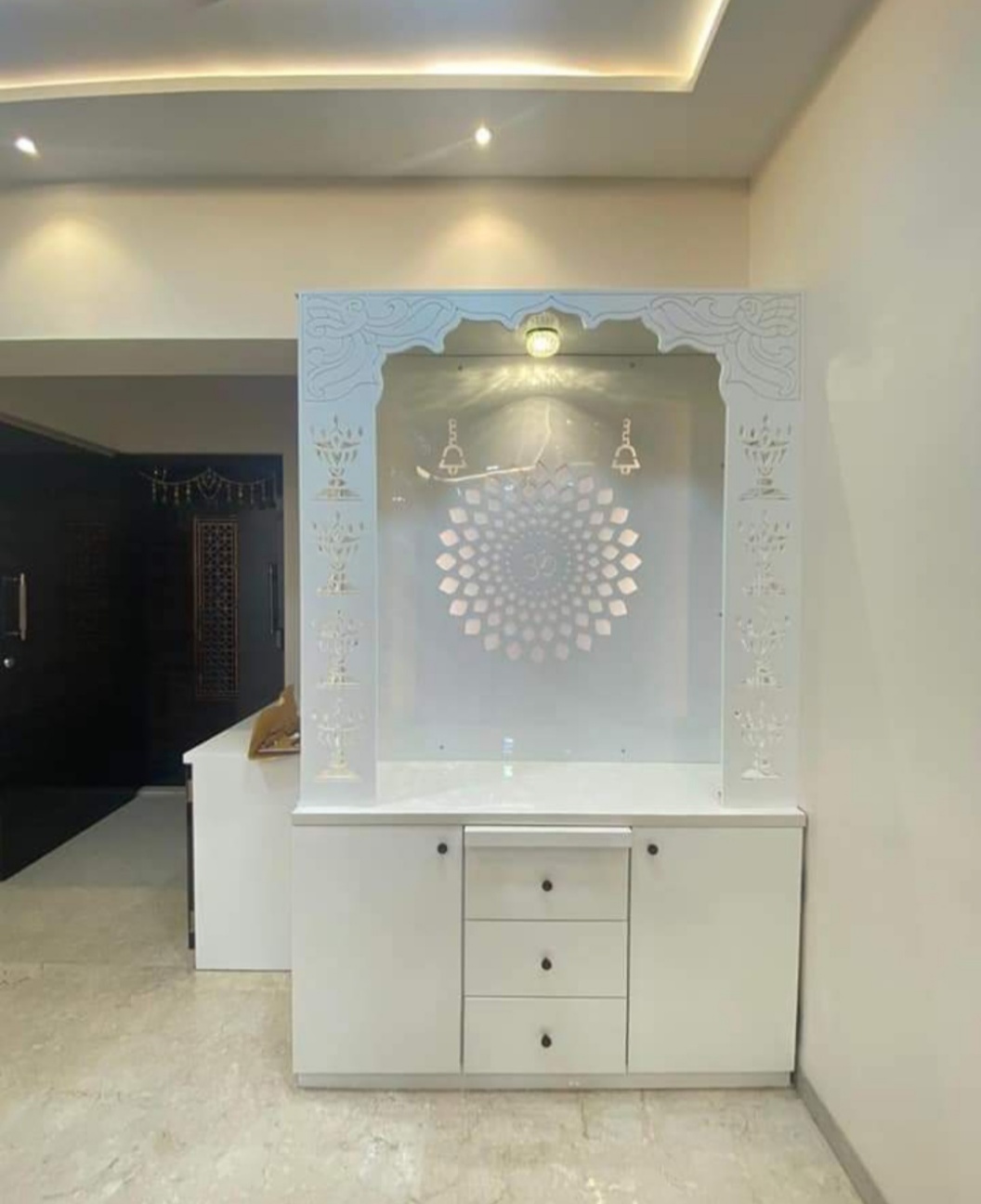 White CNC-carved pooja unit with a backlit Om lotus jaali panel and storage drawers below, set beneath a cove-lit ceiling.