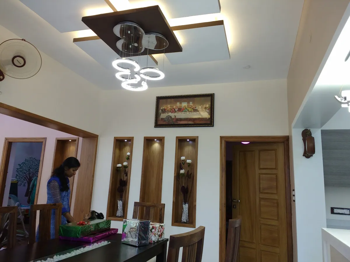Dining area of a Kerala home with three teak-framed lit wall niches holding white flower vases below a framed Last Supper print