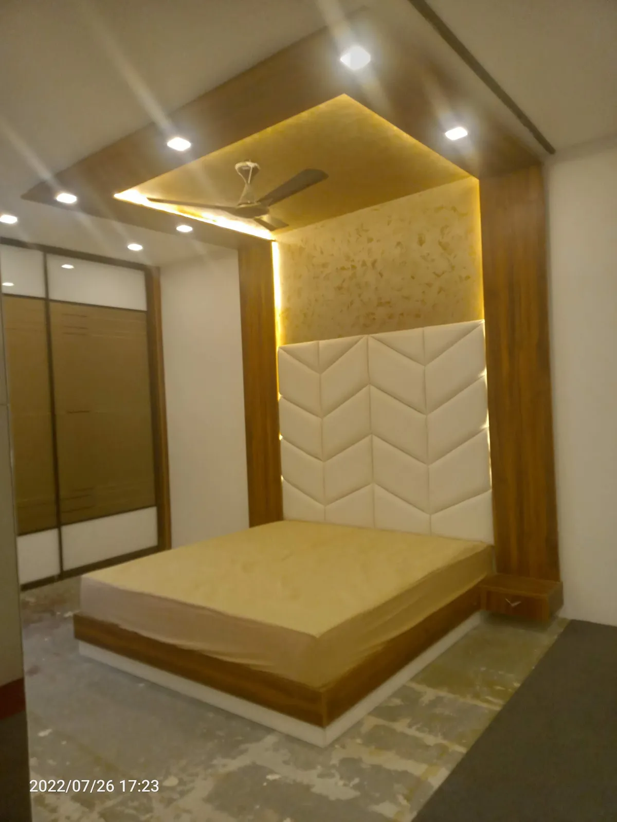 Newly finished bedroom with a chevron-quilted cream headboard set into a teak-laminate frame, a backlit gold textured panel above it
