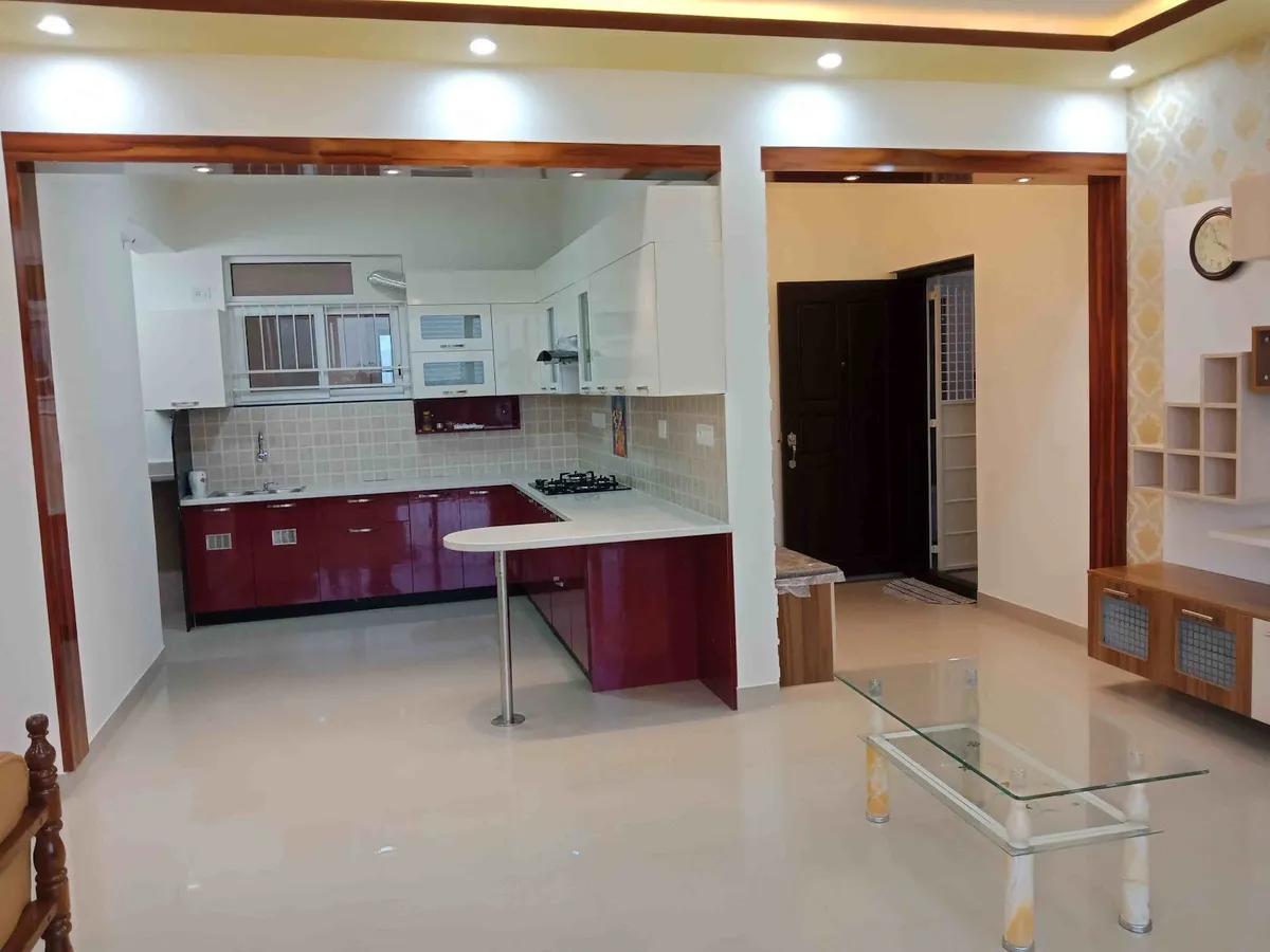 Open-plan view from an L-shaped kitchen with glossy maroon and white cabinetry and a breakfast counter, through to a living area with a wood TV unit