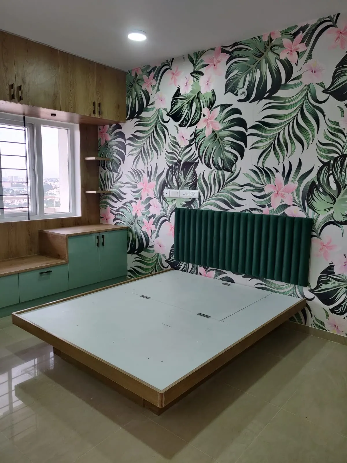 Handover shot of a bedroom with a channel-tufted emerald headboard against pink-and-green tropical wallpaper, sage and oak storage, and a bare platform bed base