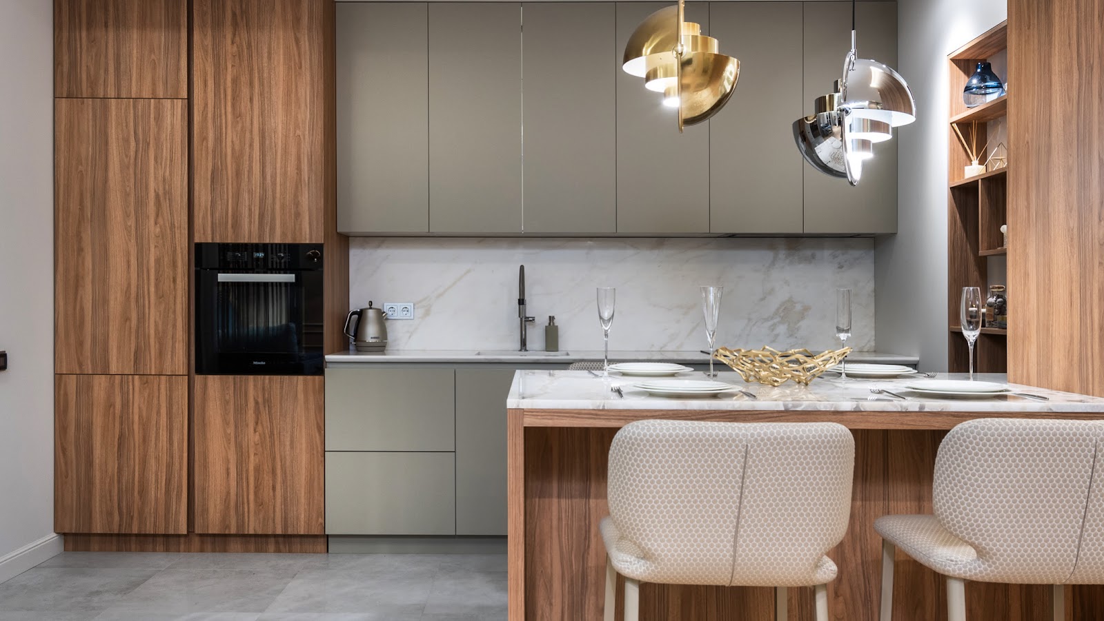 Walnut and olive-green kitchen with a marble waterfall island, gold and chrome dome pendant lights, and a built-in oven wall.