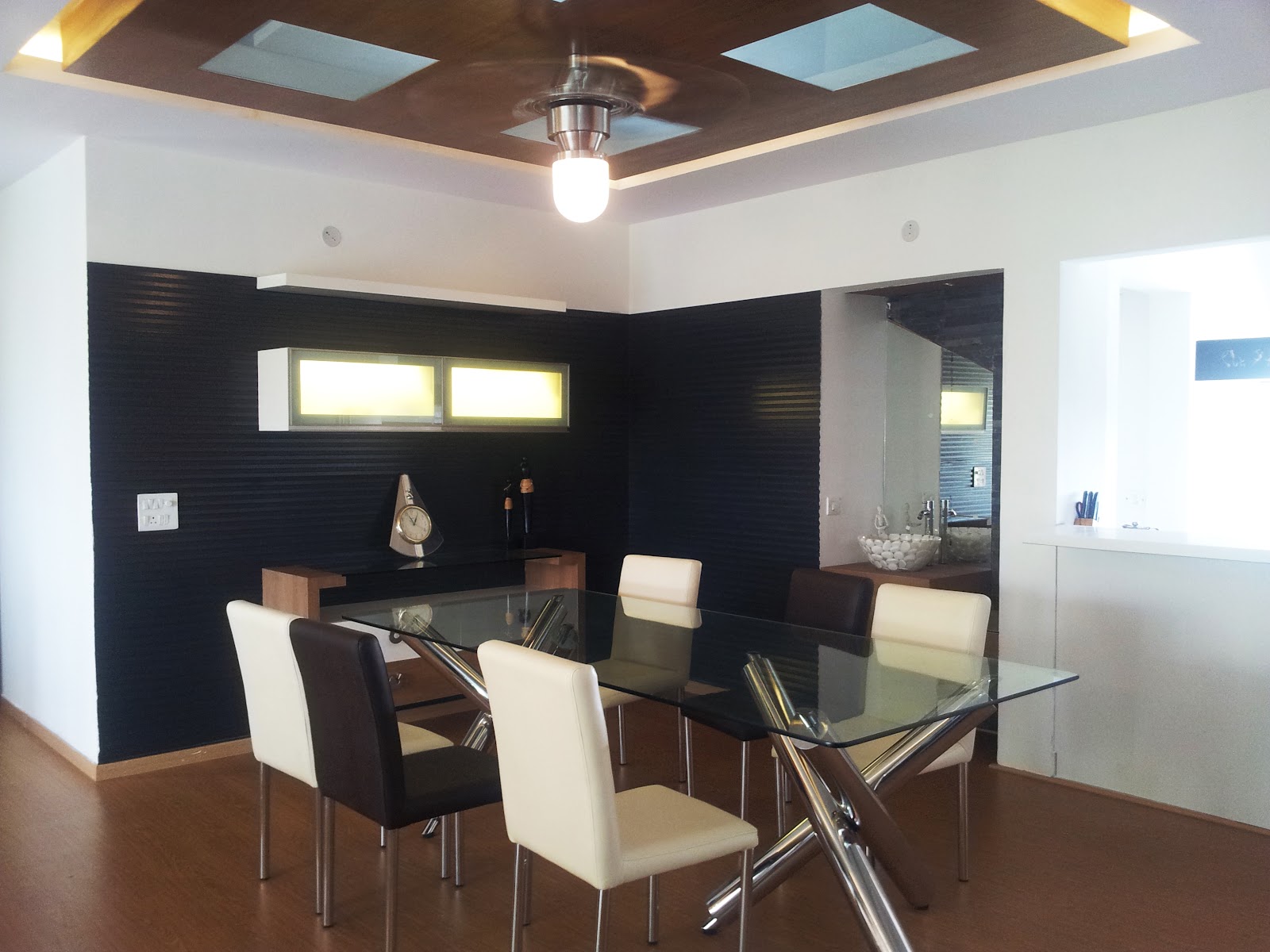 Dining area with a glass-top chrome-cross-leg table, cream and brown leather chairs, a dark ribbed accent wall with a backlit display shelf