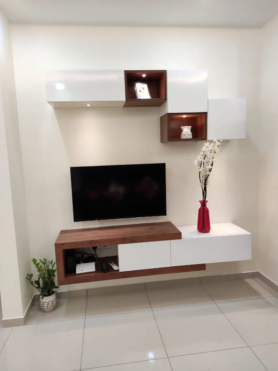 Wall-hung white-gloss and walnut TV unit with two lit open display boxes staggered above a 43-inch TV, on a bare cream wall over glossy white tile.