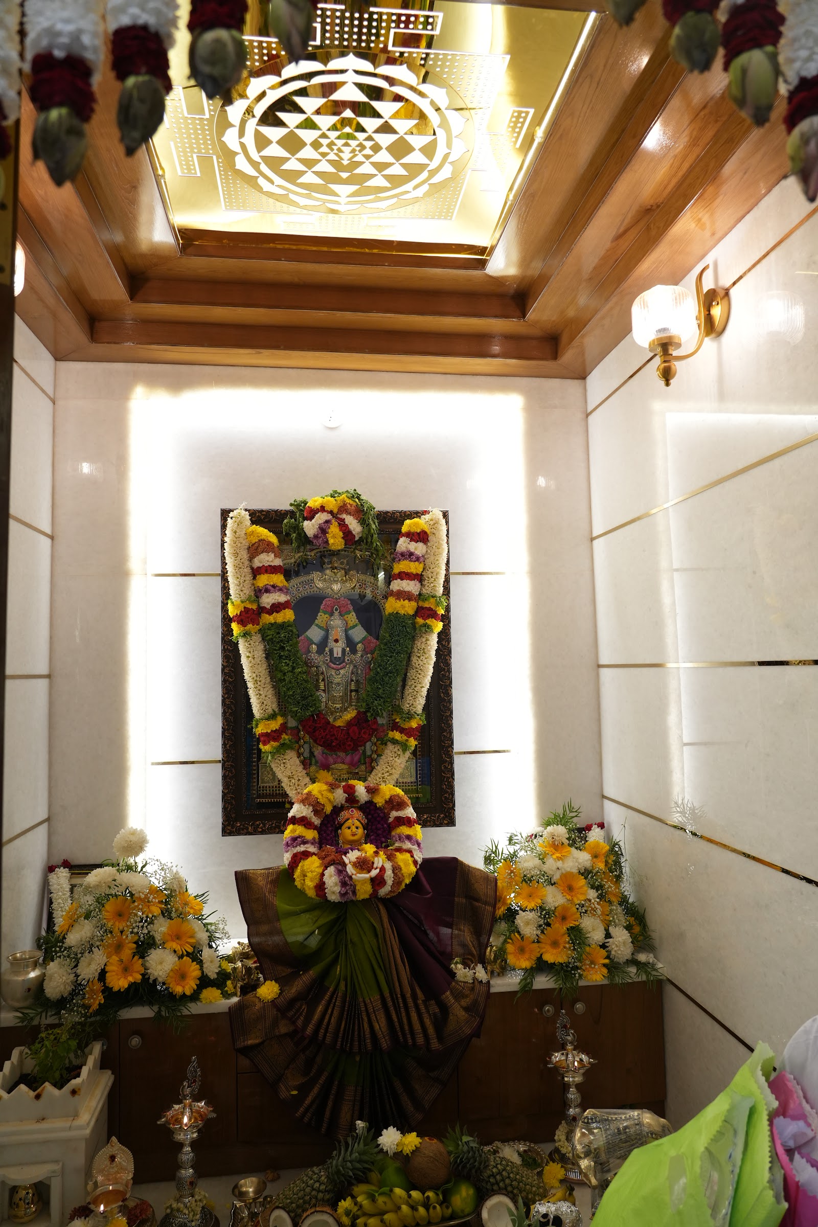 A marble-clad pooja alcove with a backlit gold Sri Yantra ceiling panel, wood ceiling trim, and a mandir framed by garlands and festive flowers