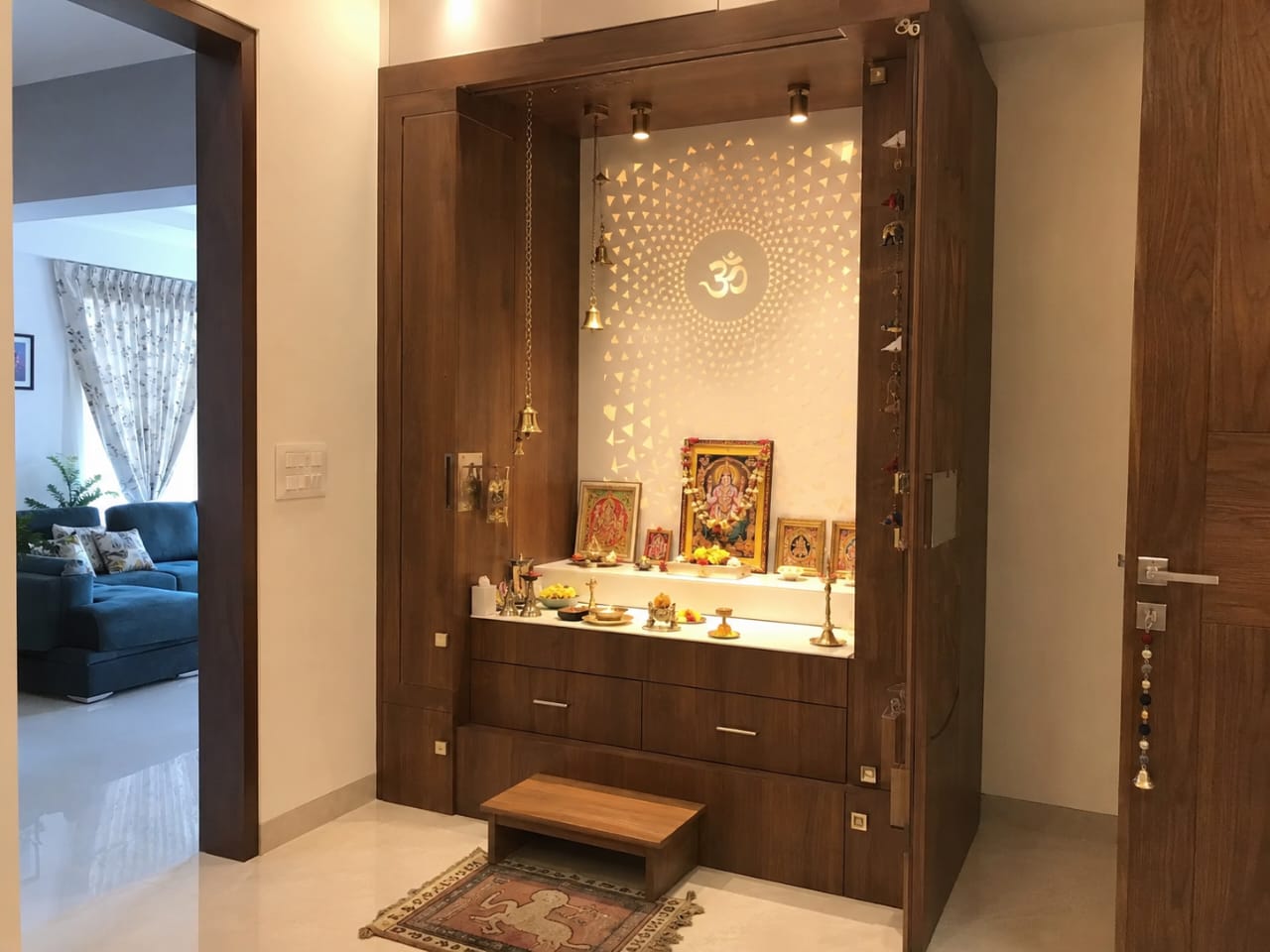 Built-in walnut-finish pooja unit with a backlit gold jaali panel featuring an Om motif, brass bells, and a stone idol shelf.