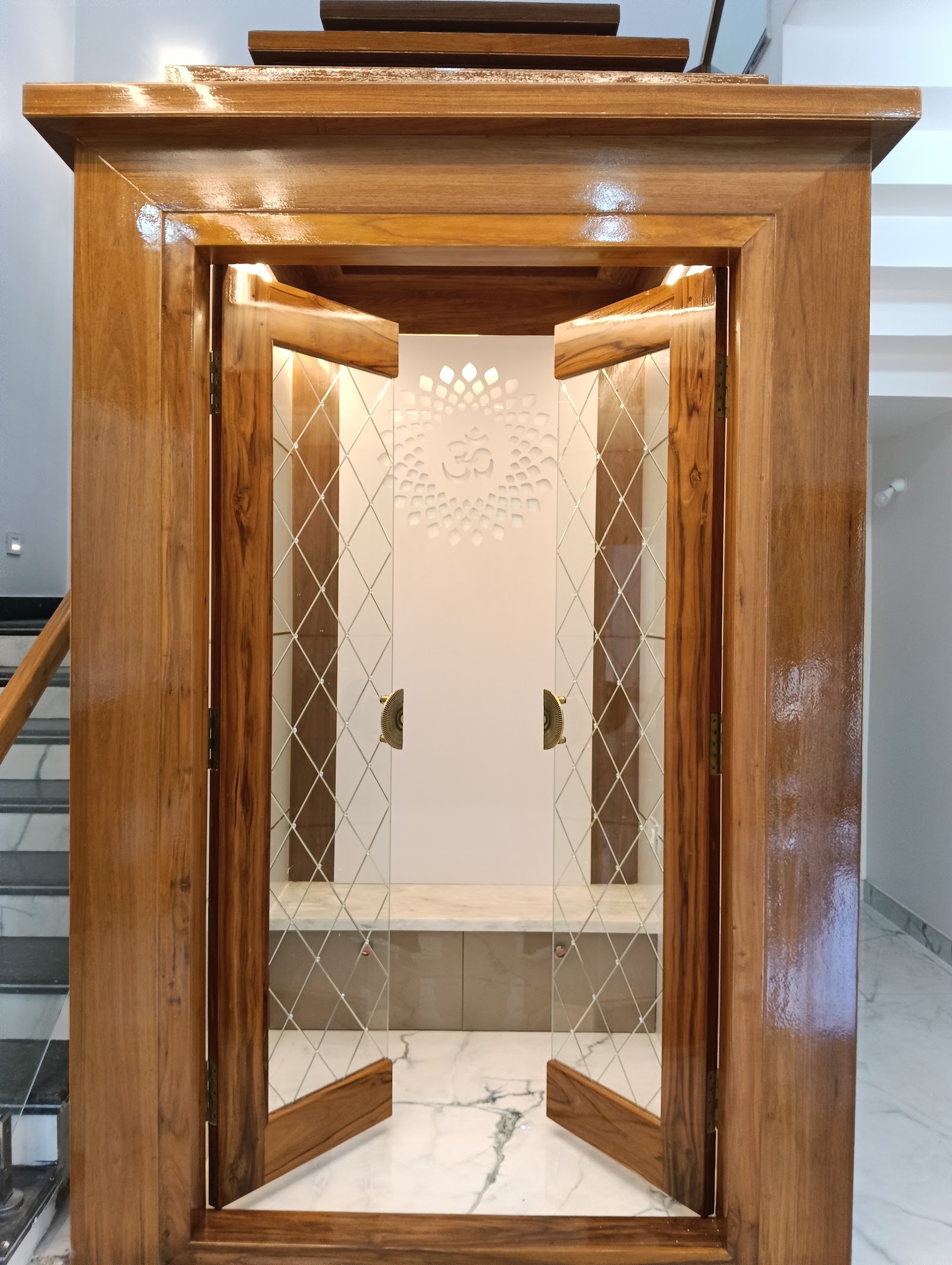 Under-stair pooja unit with glossy teak wood diamond-glass doors opening to a backlit lotus-and-Om wall motif above a marble altar shelf.