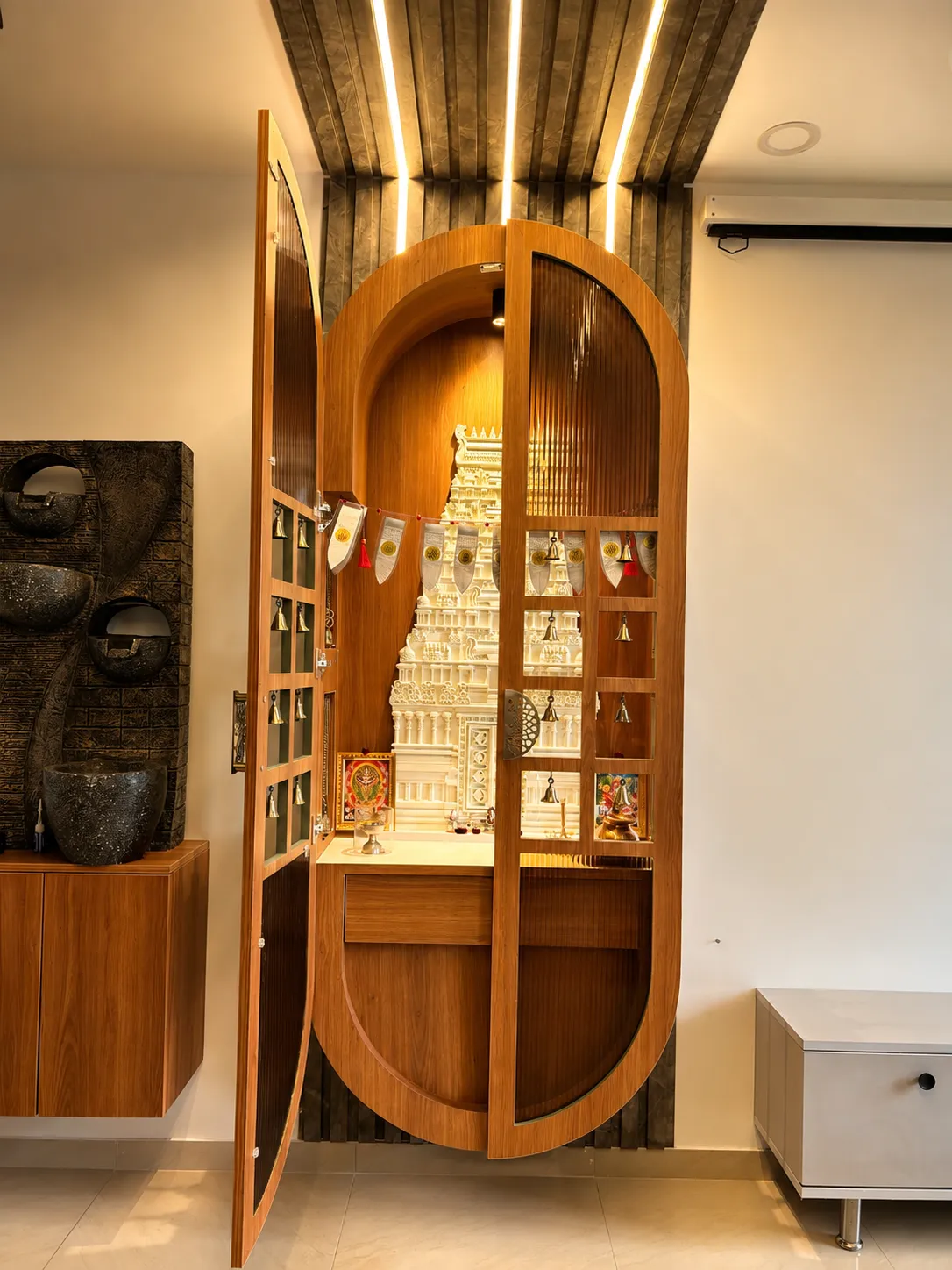 Capsule-arch teak mandir whose fluted glass doors open on rows of brass bells to a white marble gopuram in a backlit niche
