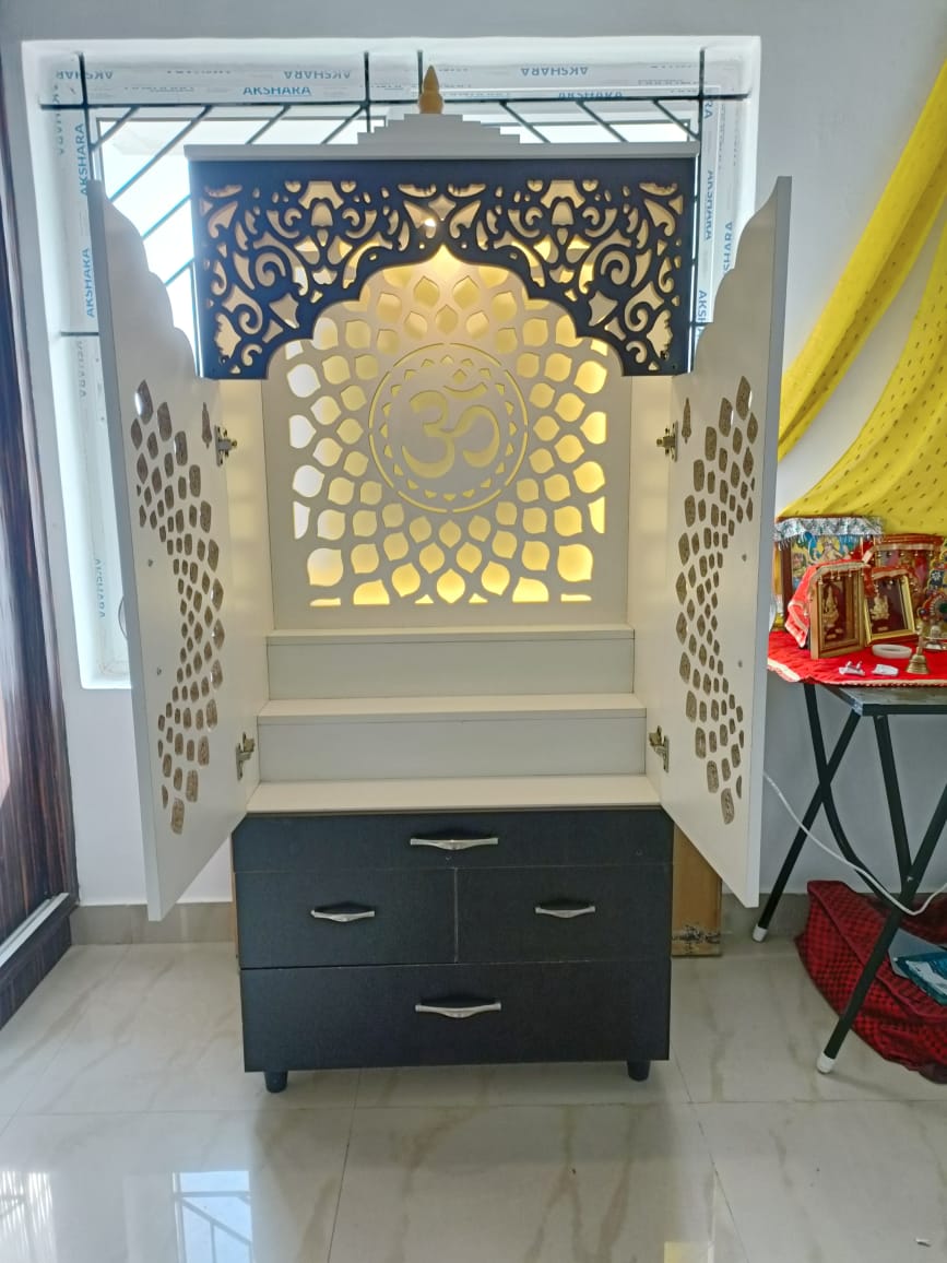 Freestanding white pooja cabinet with a backlit Om jaali panel, ornate perforated cabinet doors, and a dark drawer base