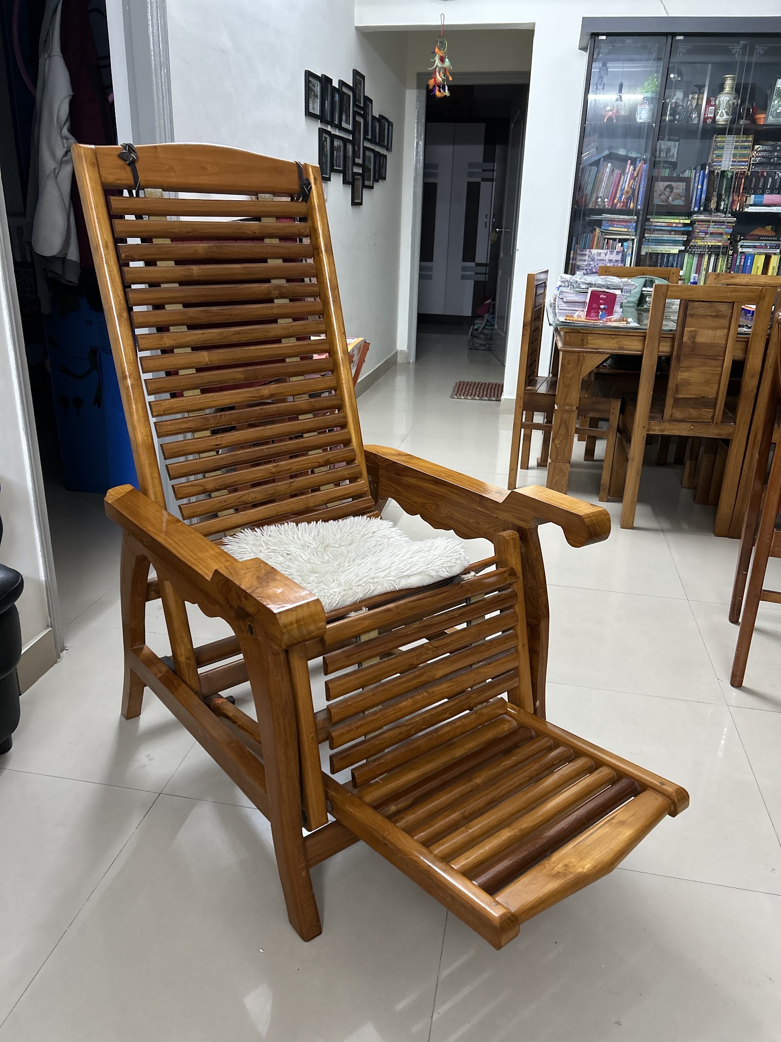 Teak wood reclining planter's chair with a fold-out leg rest in an open living-dining area, with a glass-front curio cabinet and dining table visible behind.