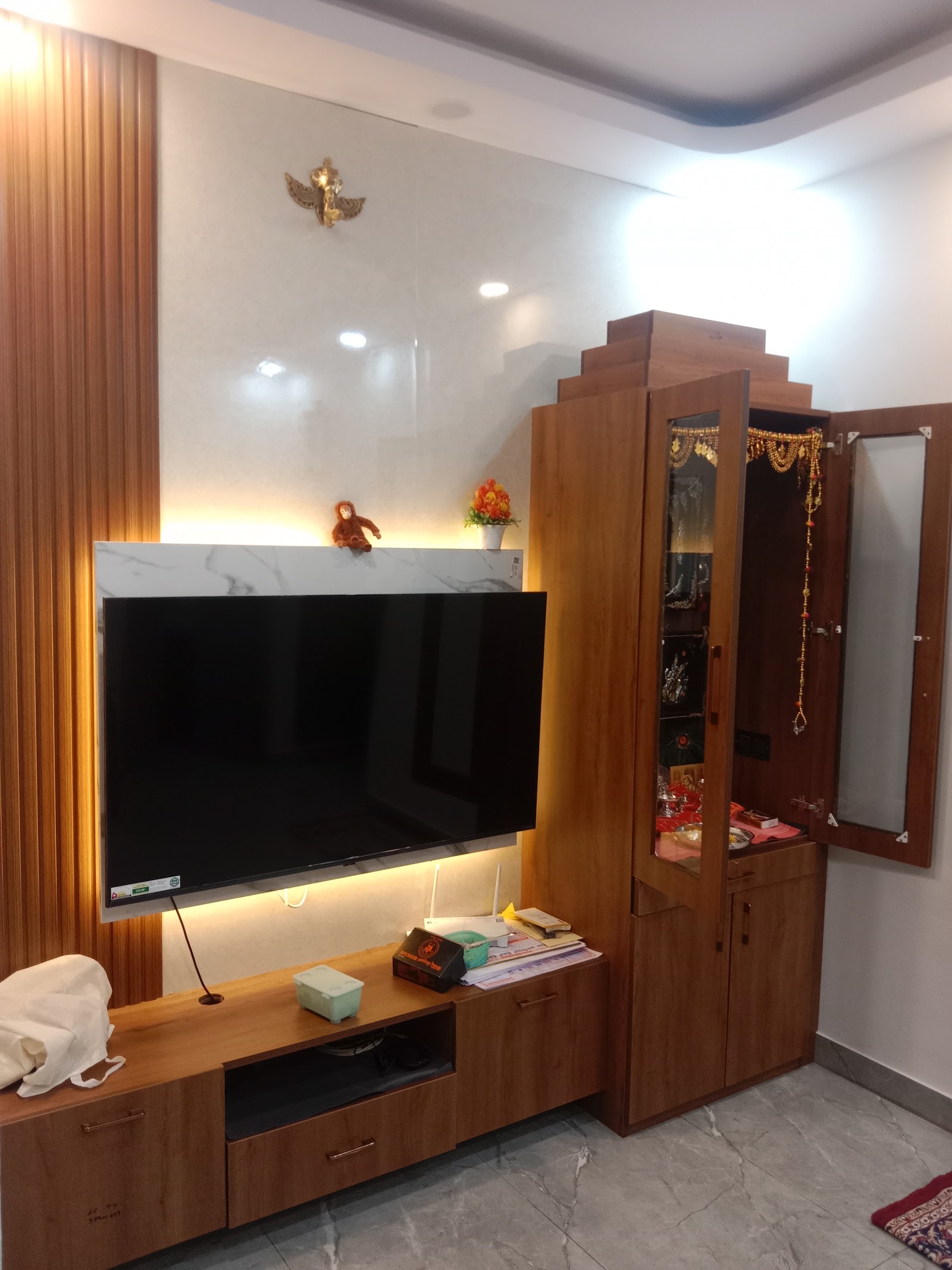 Living room TV wall with a marble-look backlit panel and an adjoining wood pooja unit with mirrored doors and temple idols.