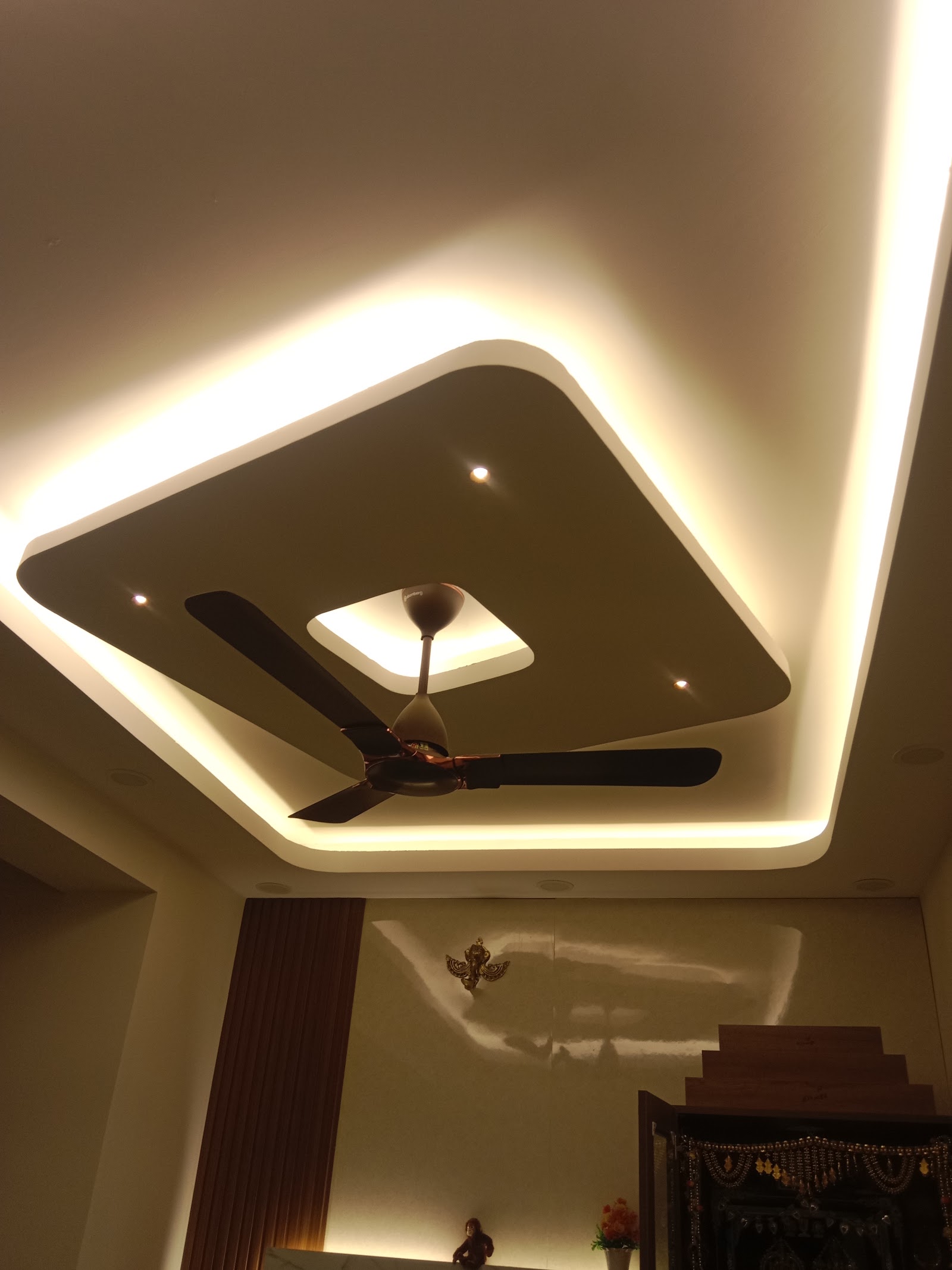Upward view of a diamond-shaped cove-lit false ceiling with a ceiling fan, above a wooden pooja mandir with beaded toran hangings visible below.
