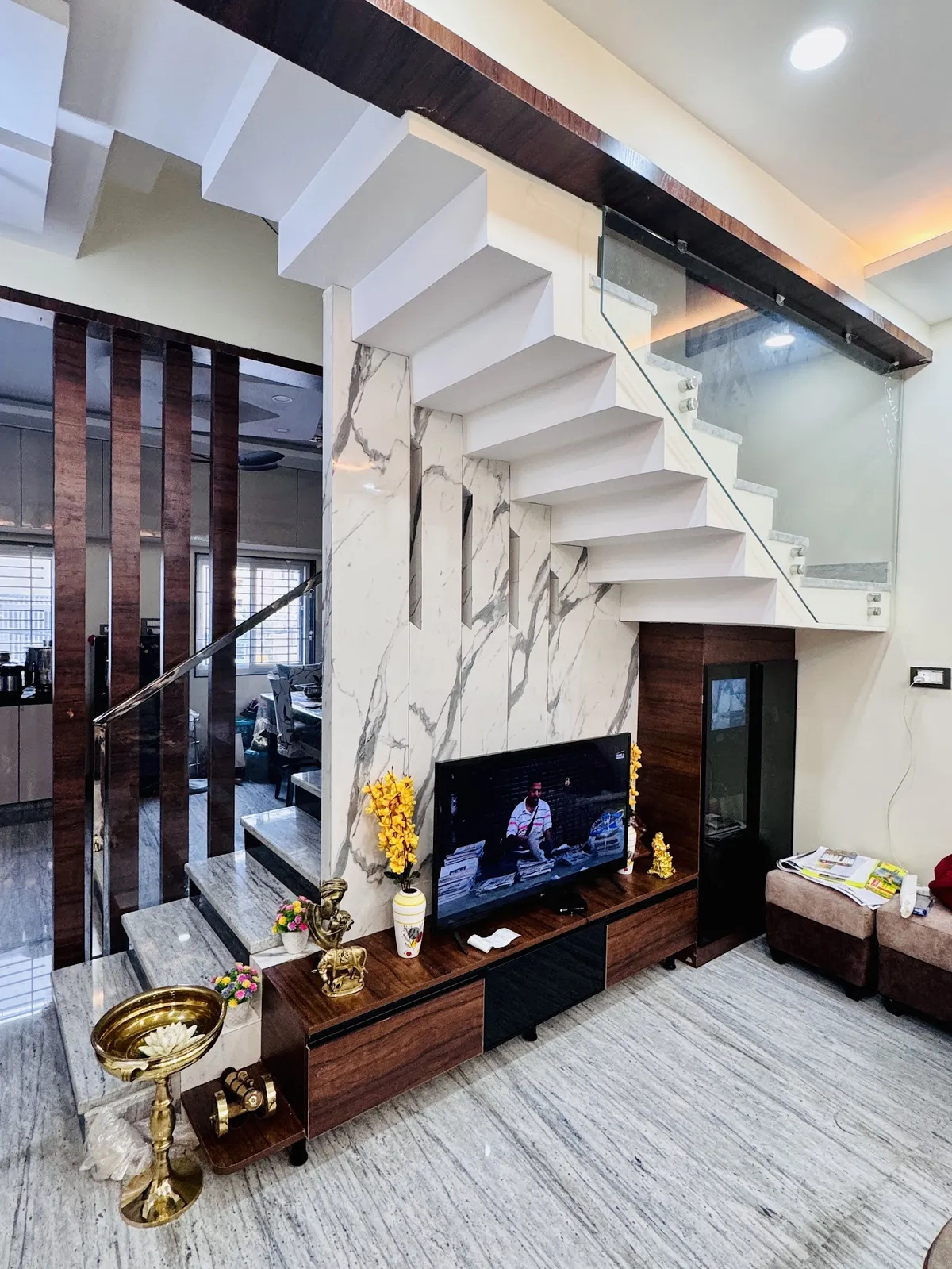 Living area tucked under a white cantilevered staircase with a glass balustrade, TV on a walnut low unit against a book-matched white-and-grey marble slab