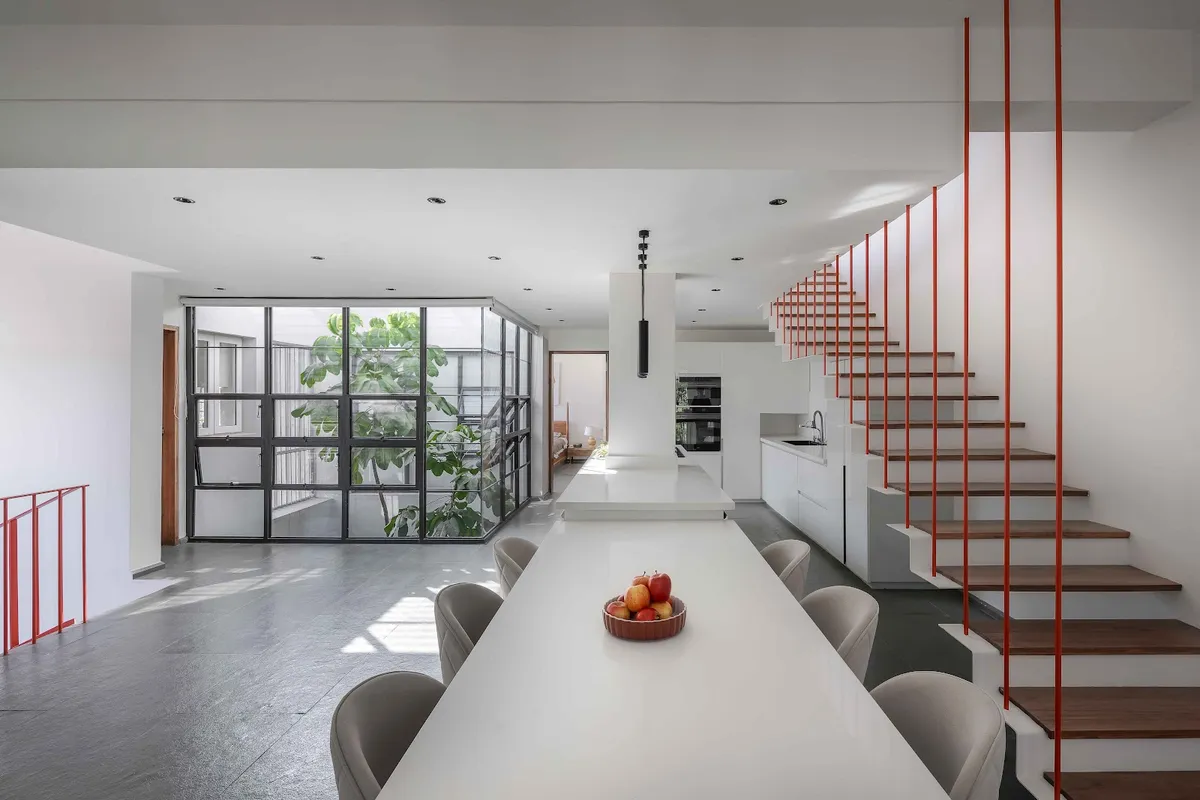 An open-plan kitchen and dining space with a long white island-dining table, built-in wall ovens, a black steel-framed glass wall to an internal courtyard