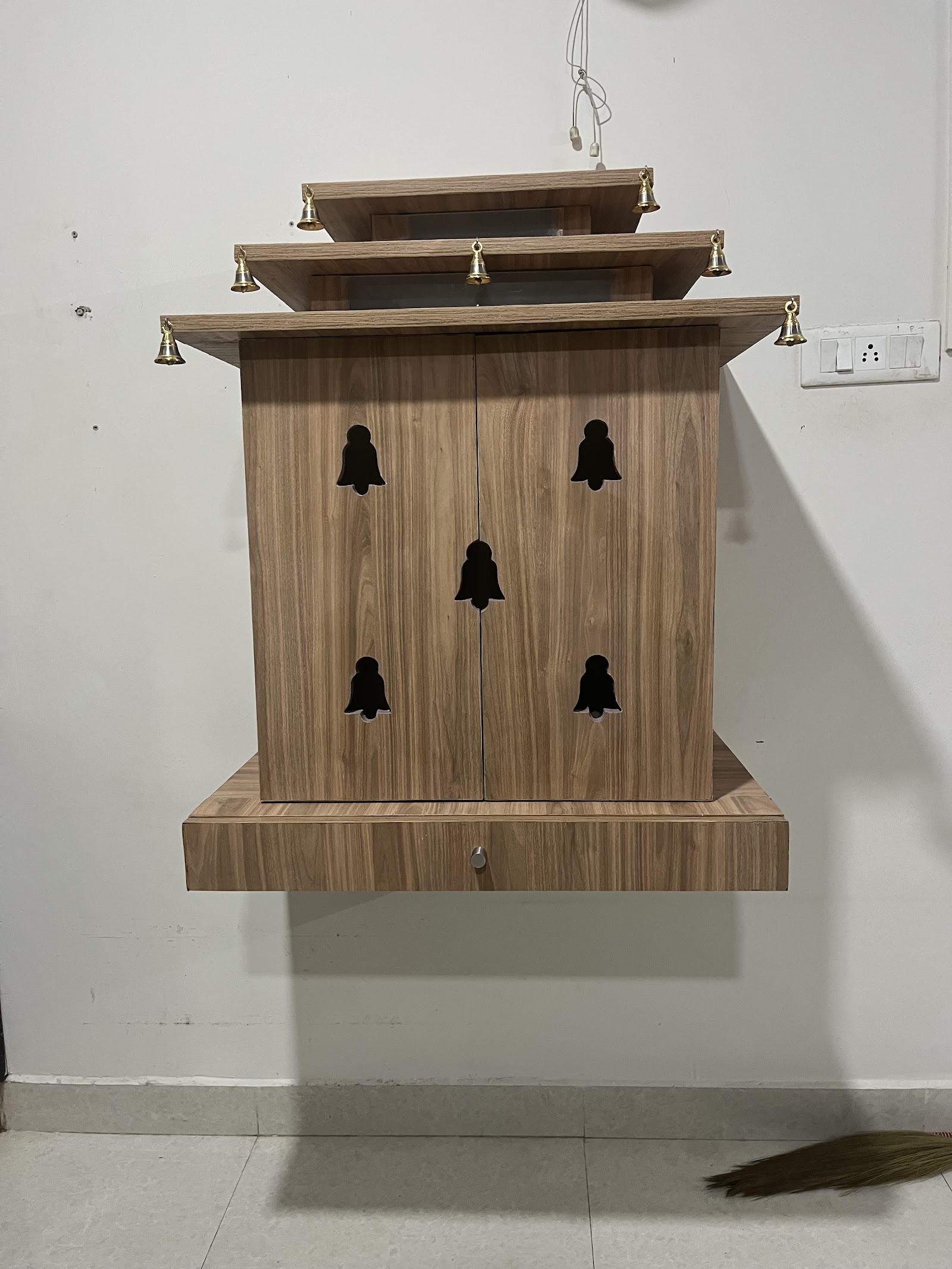 Wall-mounted wooden pooja unit with a tiered canopy roof, bell-motif cutout doors, and hanging brass bells.