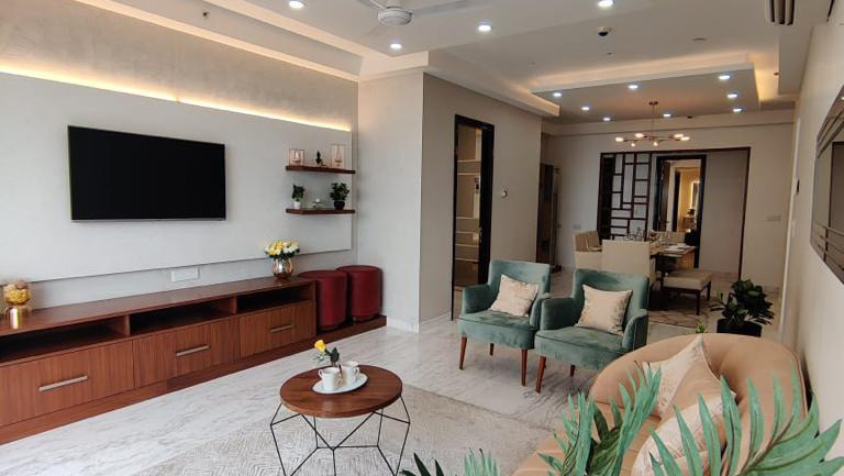 An open living area with a wood-veneer TV console under cove lighting, teal velvet armchairs around a wire-frame coffee table