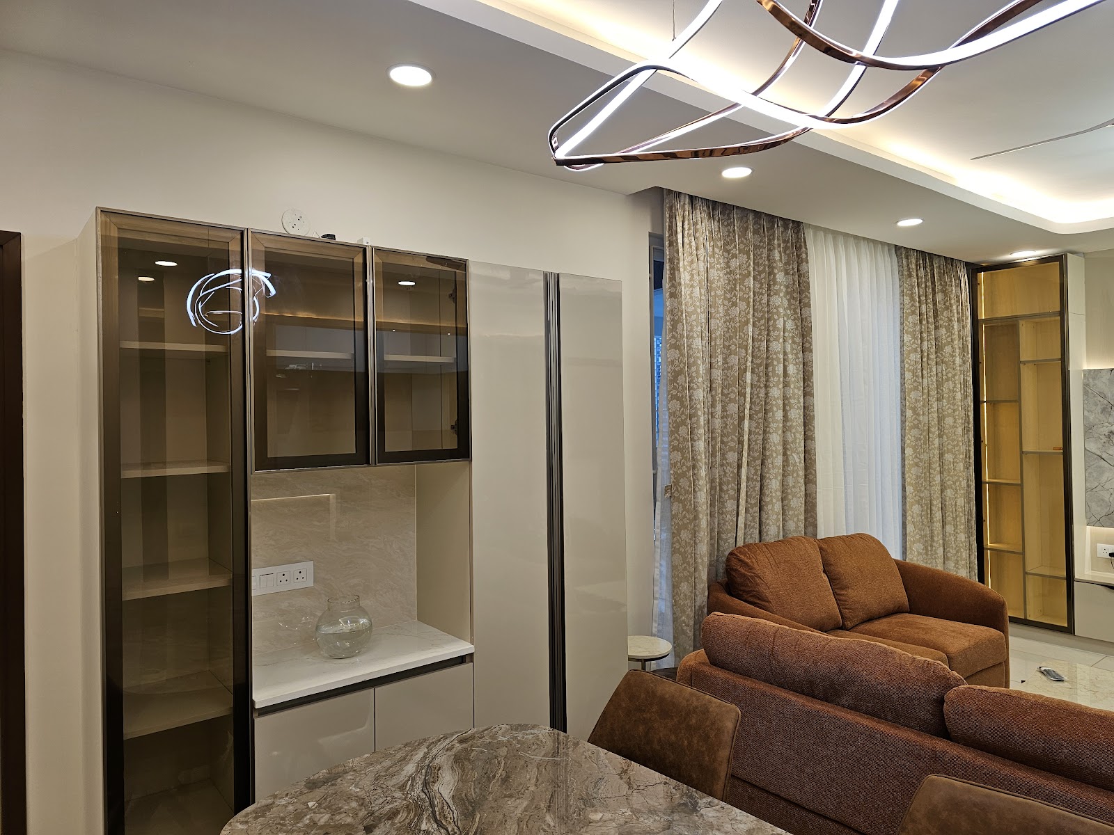 Open-plan living space with a mirrored glass-front display cabinet, floral curtains, a sculptural ribbon pendant light