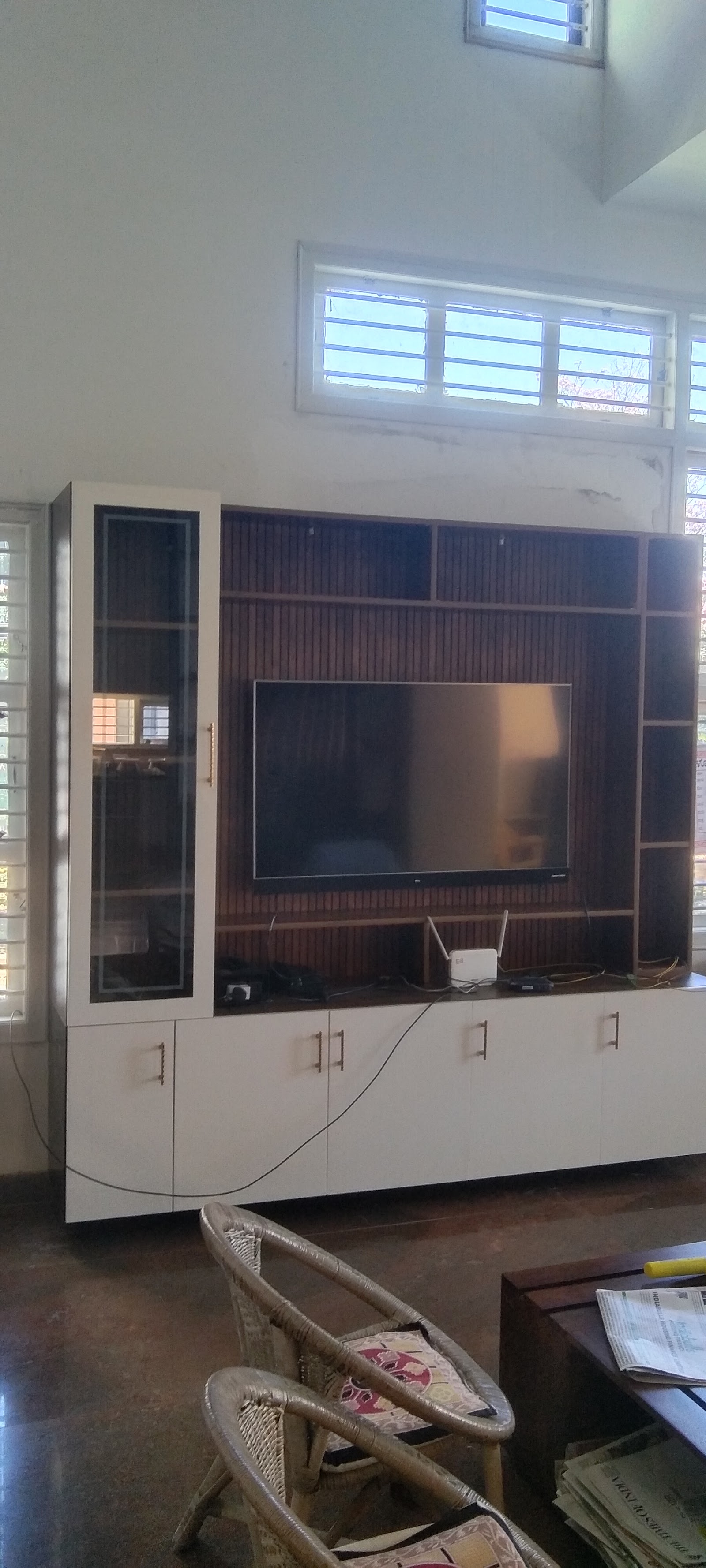 Living room TV wall unit with a wood-slat backdrop and glass-front display cabinet, white base cabinets, facing a pair of wicker chairs.